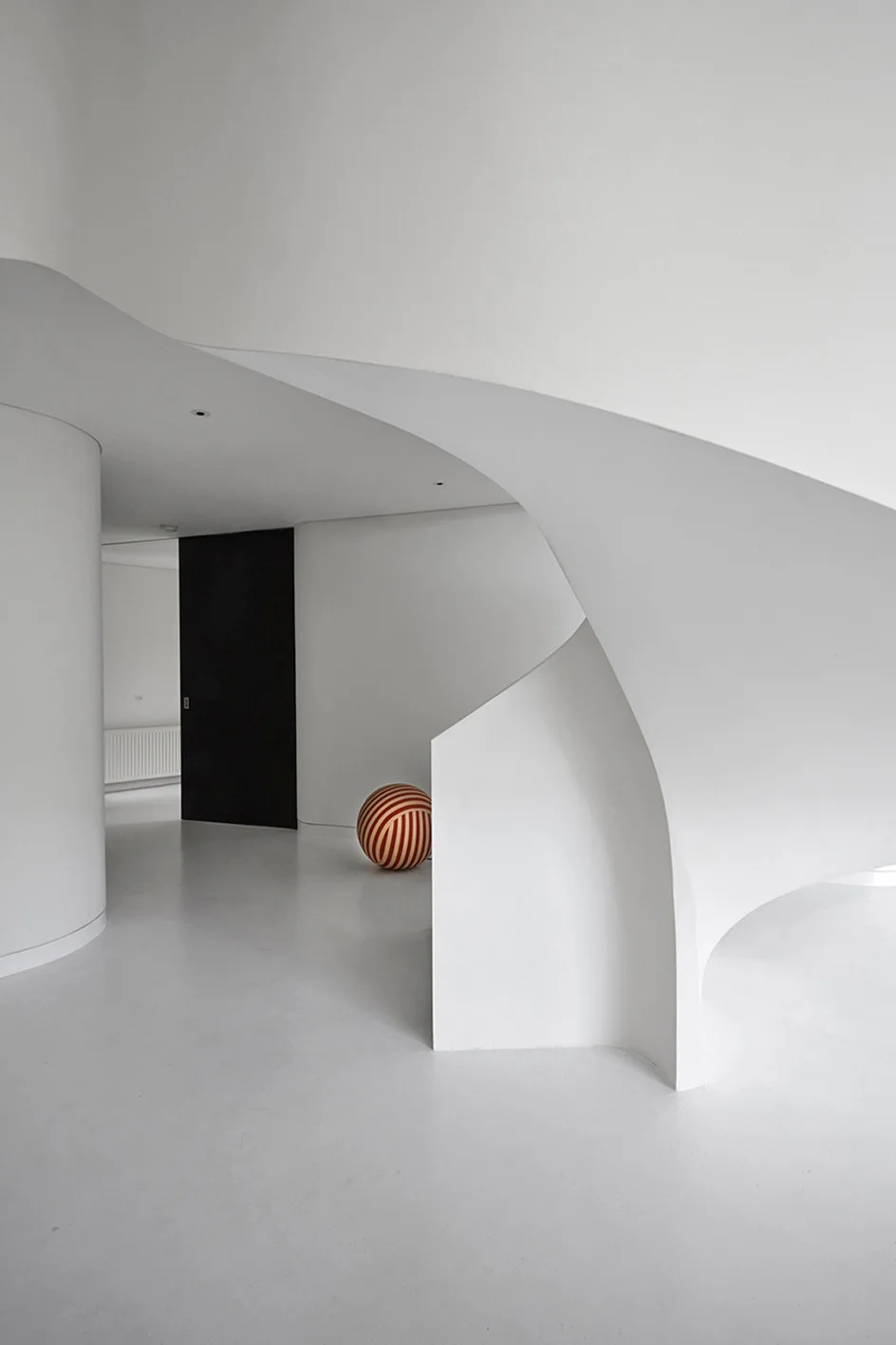 A minimalist white hallway featuring a sculptural curved staircase, seamless floors, a black door, and a red and white striped sphere on the ground.