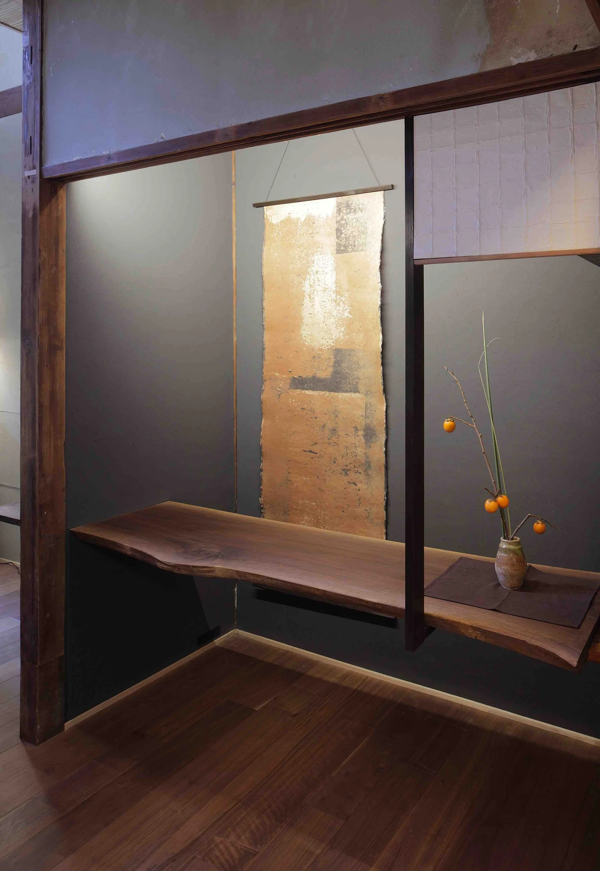 A minimalist dark grey alcove featuring a floating live-edge wood shelf, a gold wall scroll, and a ceramic vase with an orange-fruited branch.