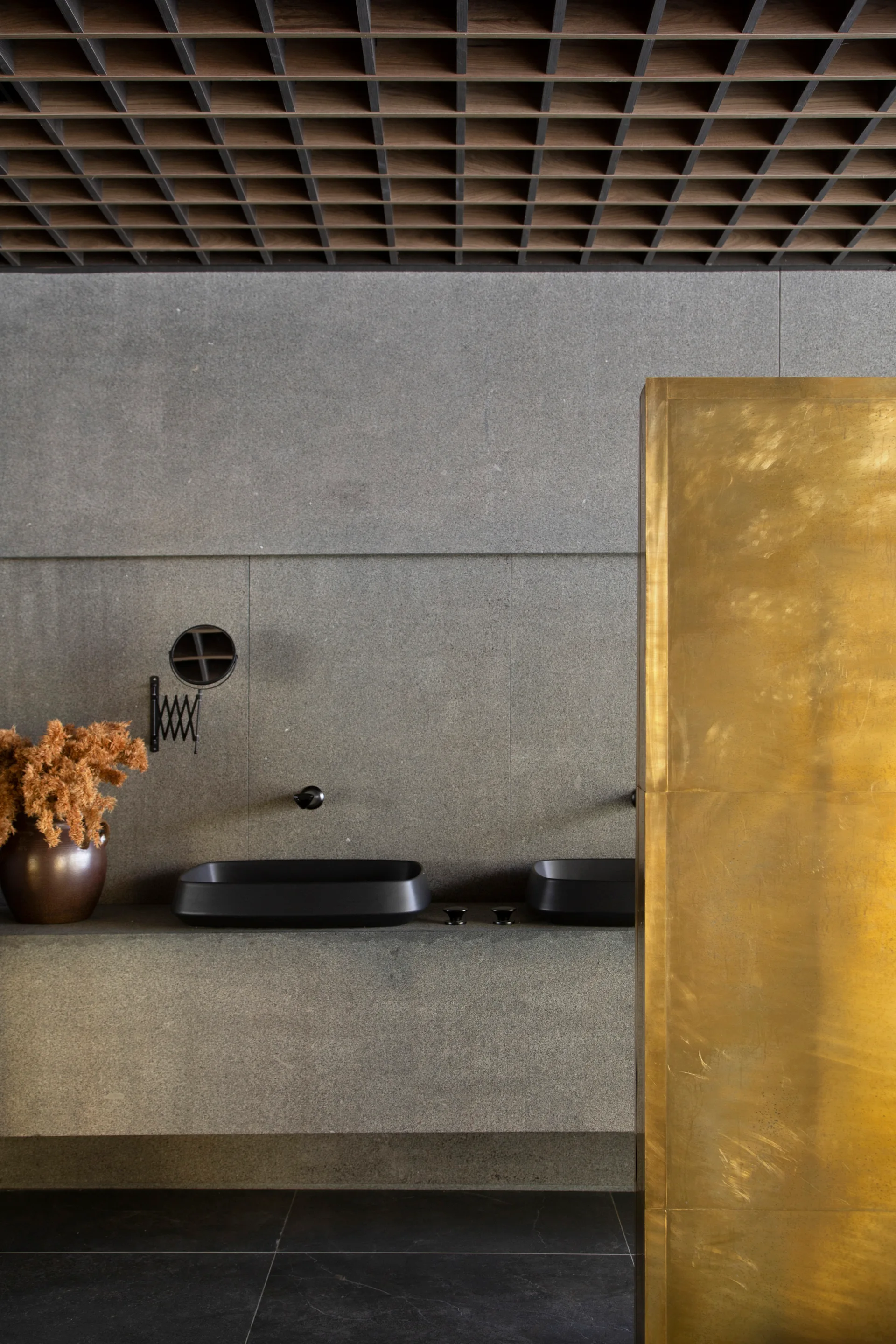A minimalist bathroom vanity features dual matte black basin sinks on a stone countertop beneath a wooden grid ceiling and next to a tall brass partition.