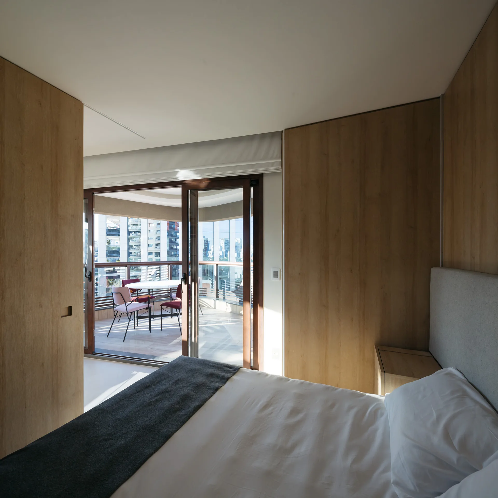 A modern bedroom featuring wood-paneled walls, a bed with white bedding, and sliding glass doors that open to a sunlit outdoor balcony.