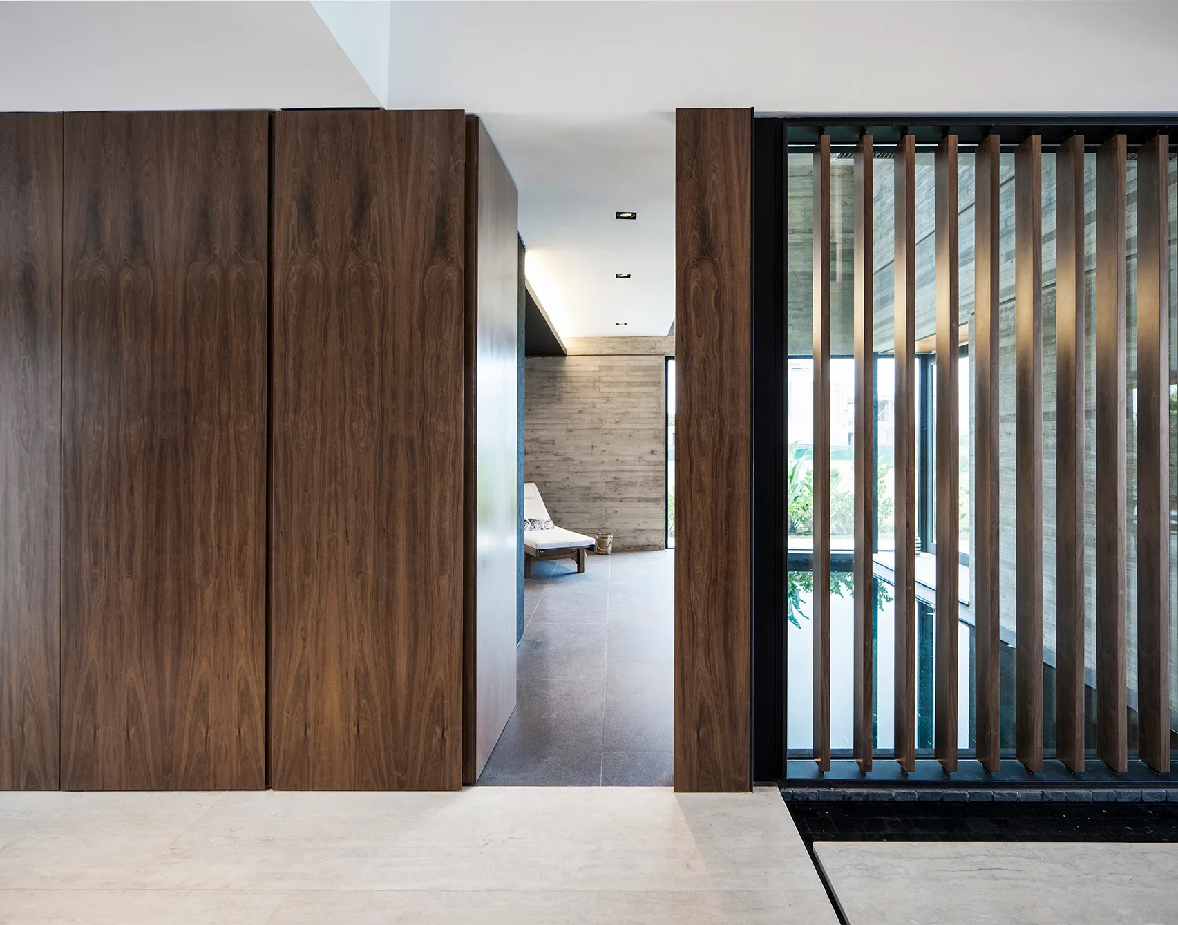 An interior corridor features large dark wood wall panels, grey tile flooring, and a vertical wood slat screen overlooking a water feature.
