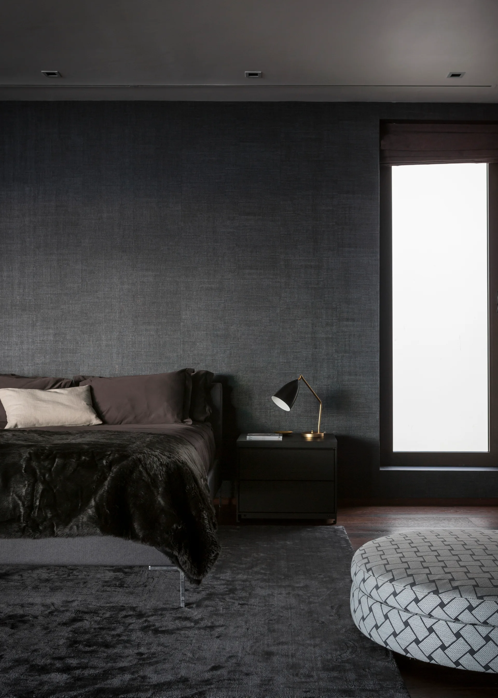 A dark bedroom features textured charcoal wallpaper, an upholstered bed with a faux fur throw, a black nightstand, and a patterned round ottoman.