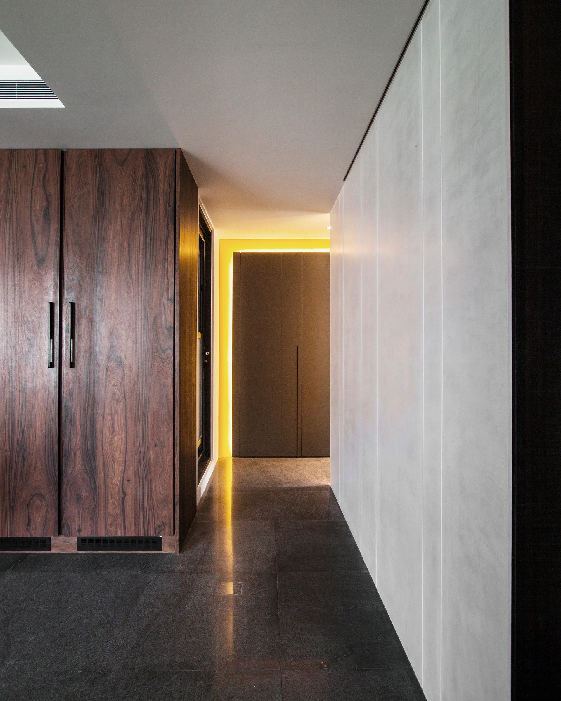 A minimalist corridor featuring built-in dark wood veneer cabinets, light grey wall panels, and warm yellow cove lighting at the far end.