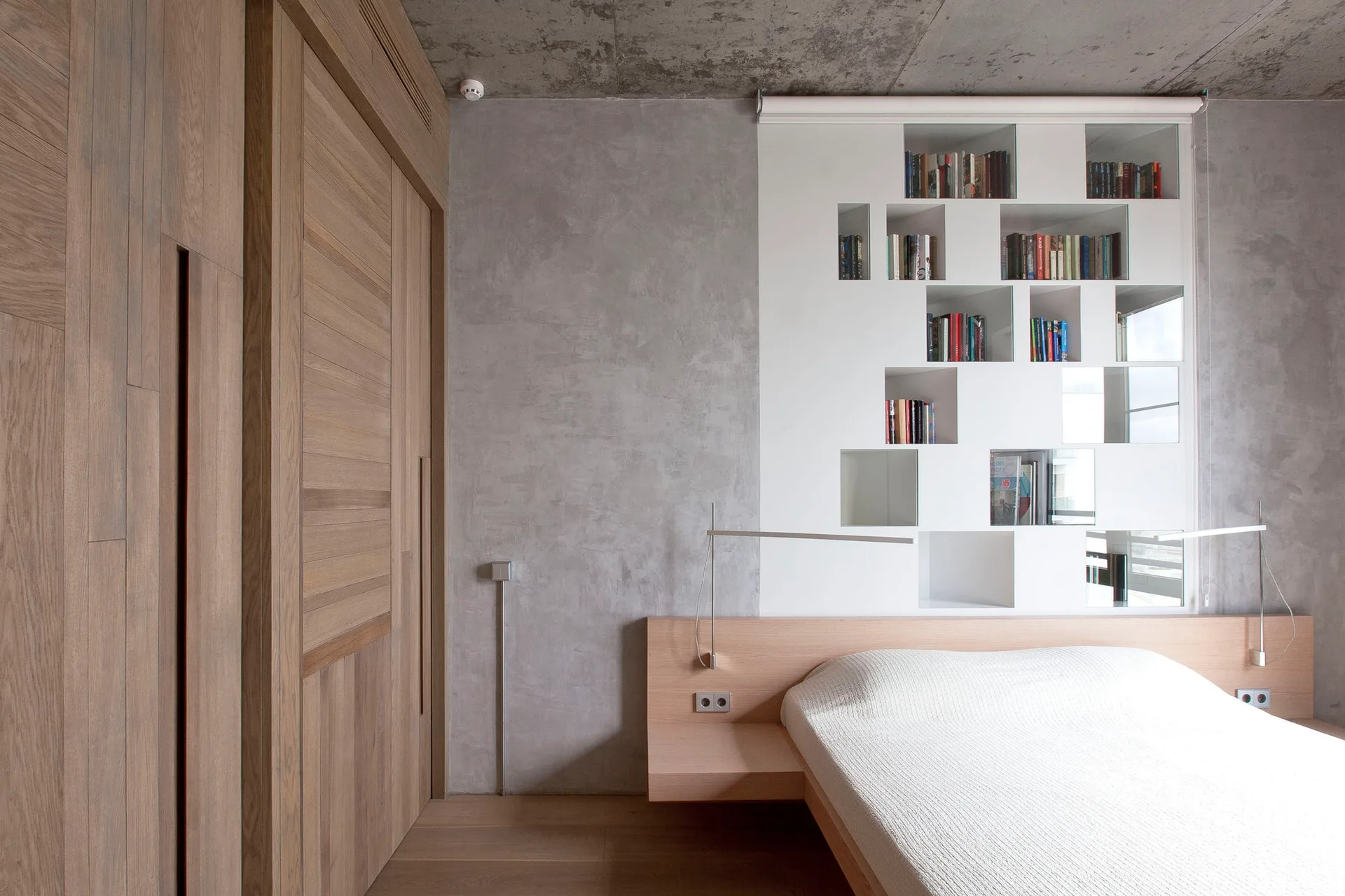A modern bedroom features a low wood platform bed, an integrated white grid shelving unit on a grey plaster wall, and a raw concrete ceiling.