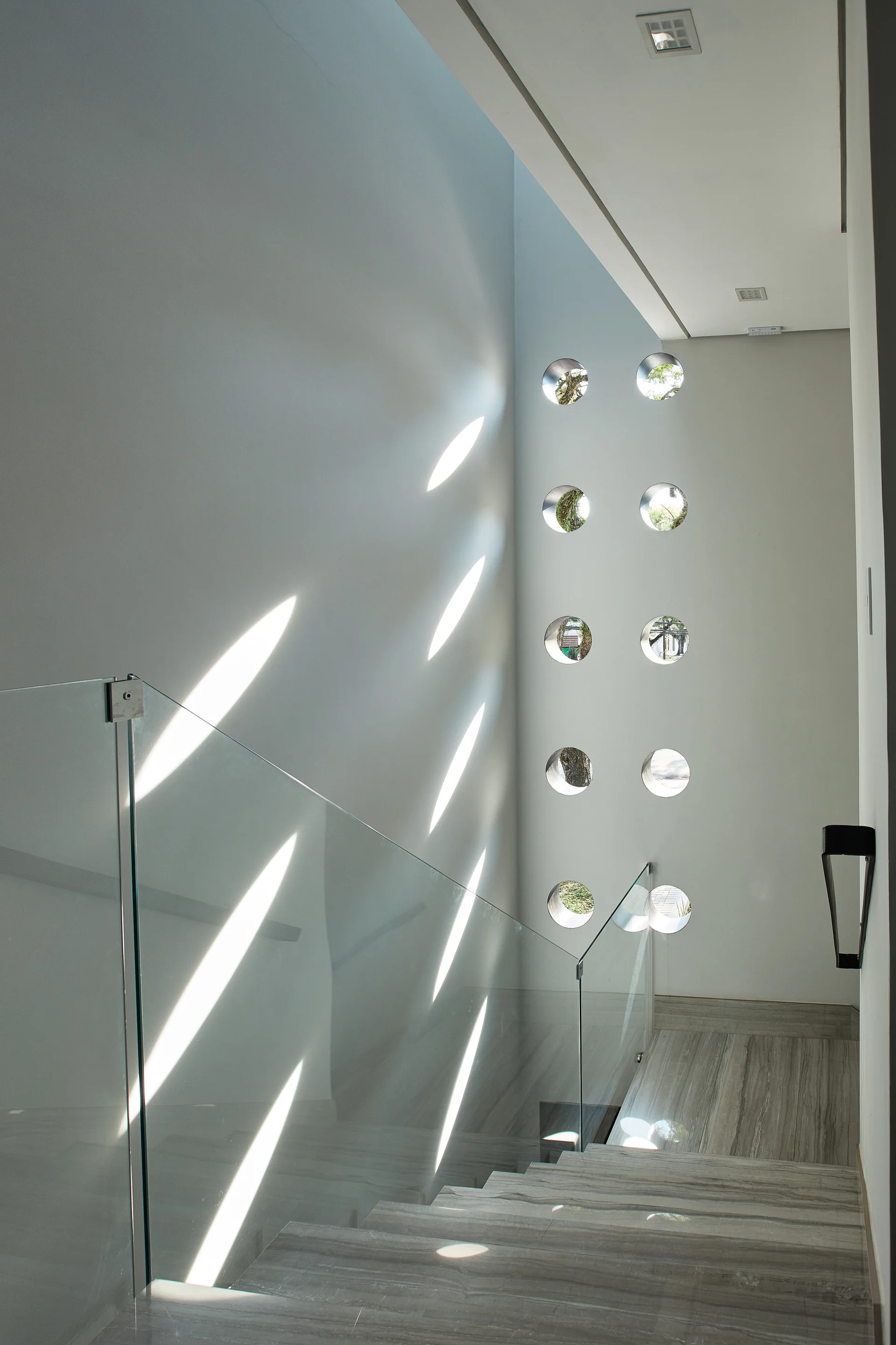 A grey stone staircase with a clear glass railing sits next to a white wall with circular windows casting bright light patterns.