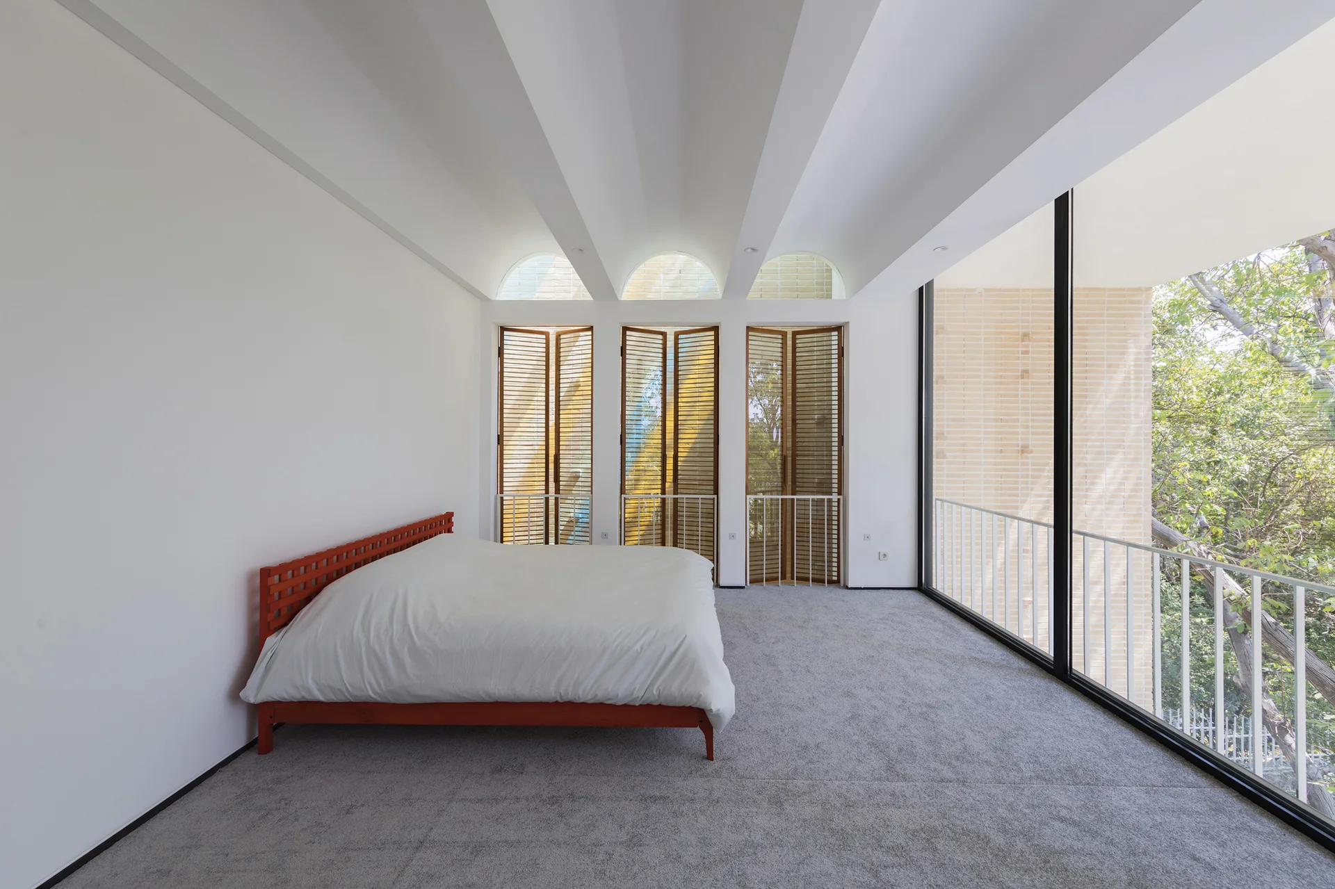 A minimalist bedroom features a red wooden bed frame, three arched windows with wood shutters, and a large sliding glass door opening to a balcony.