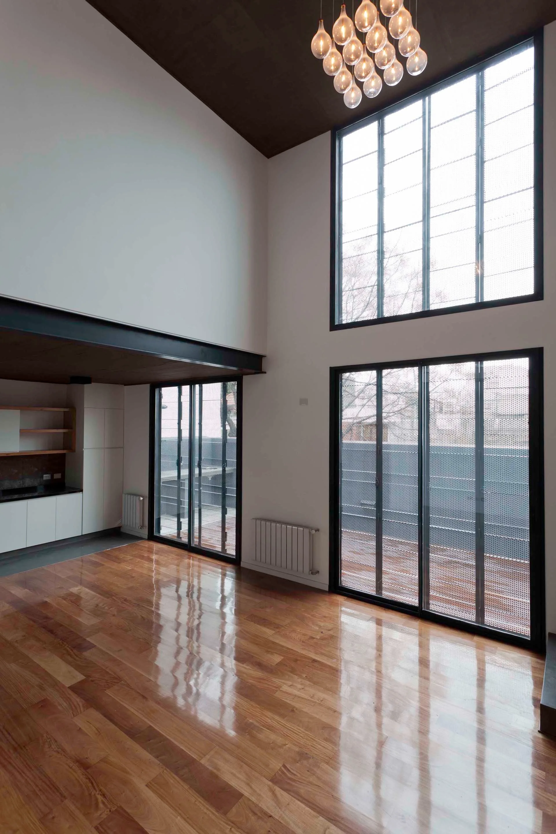 A double-height modern room features polished hardwood flooring, tall black-framed glass windows, and a glass globe pendant light cluster.