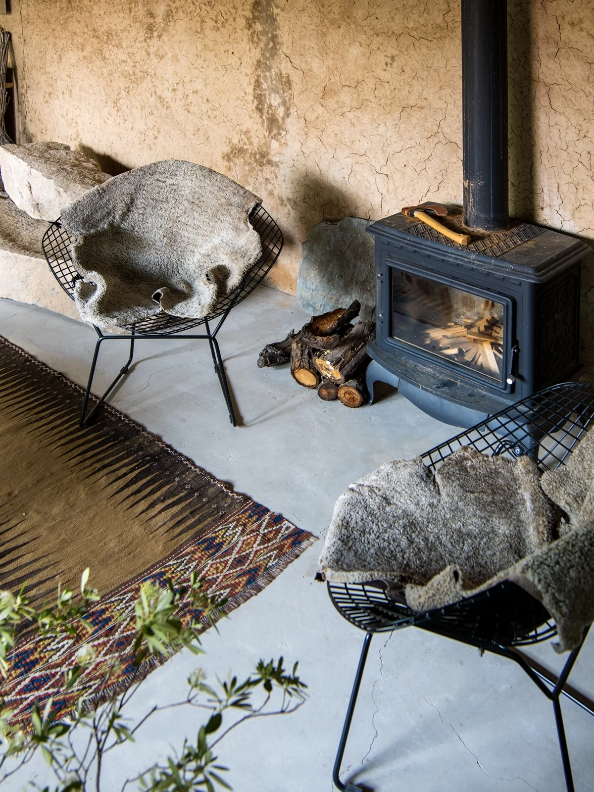 Two black metal wire chairs with shaggy fur throws face a black wood-burning stove against a textured tan plaster wall.