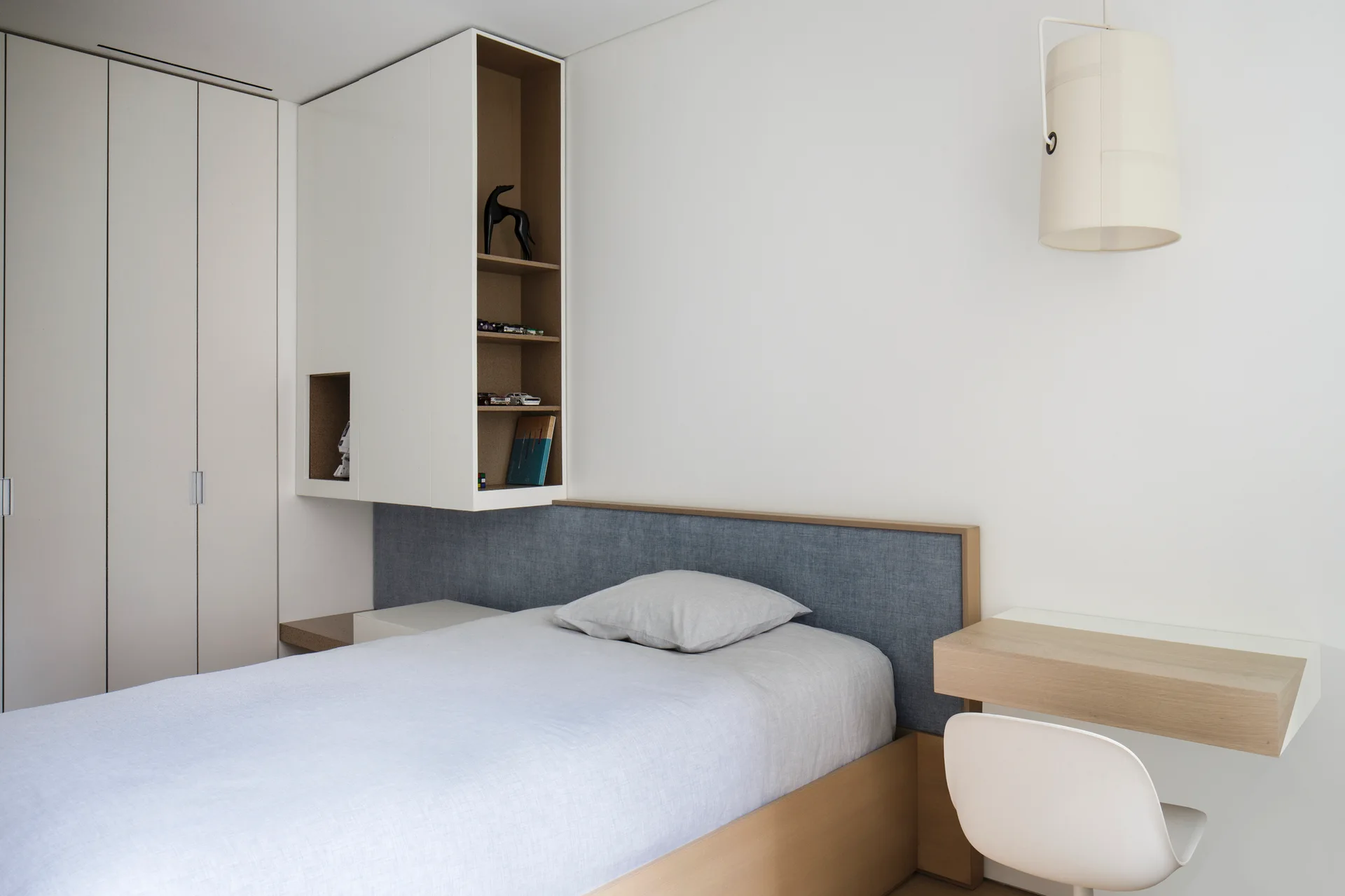 A minimalist bedroom featuring a single bed with a grey headboard, custom white wall cabinets, and a wall-mounted wood desk with a white chair.