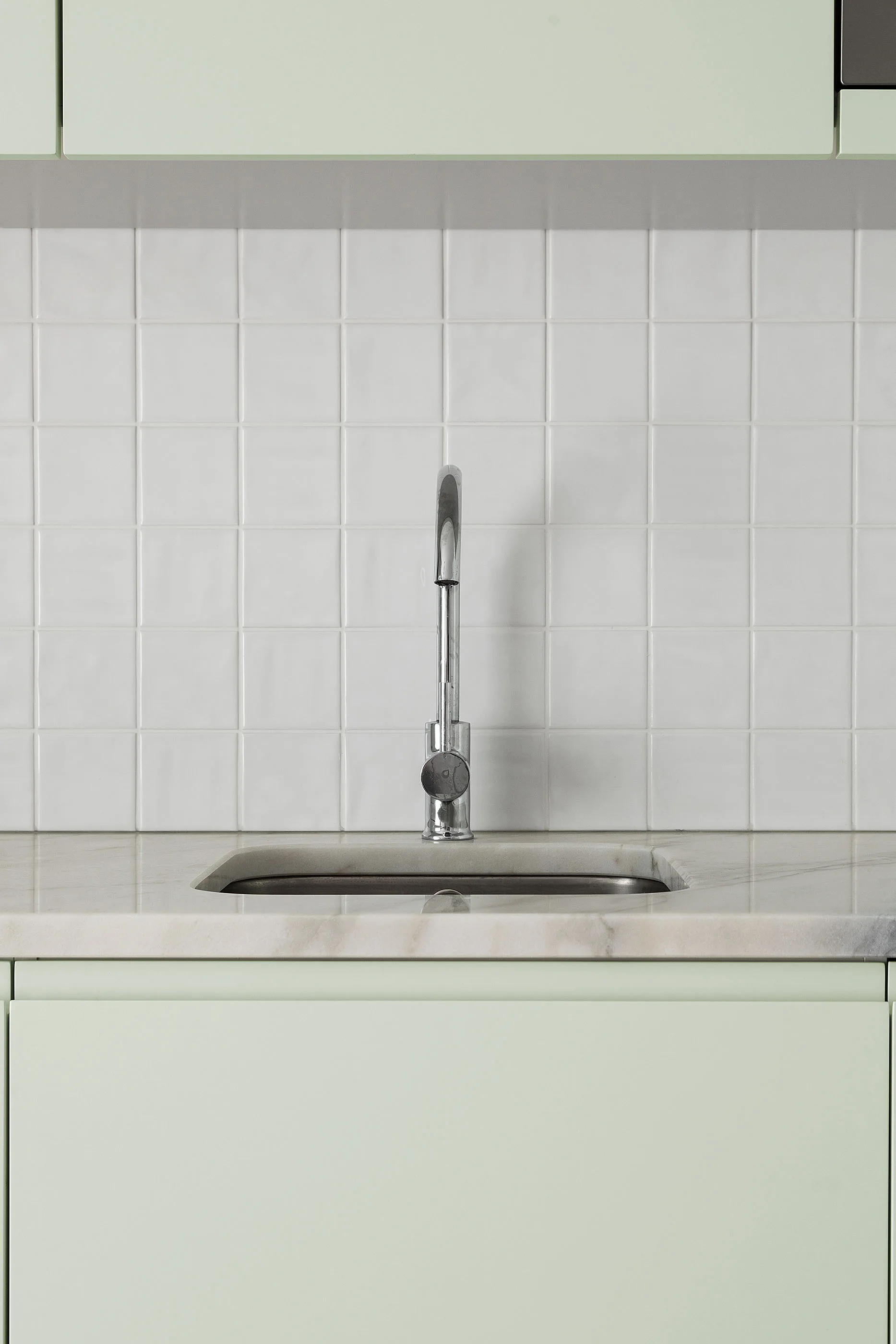 A close-up of a kitchen sink and faucet set in a marble countertop, flanked by mint green cabinets and a white square tile backsplash.