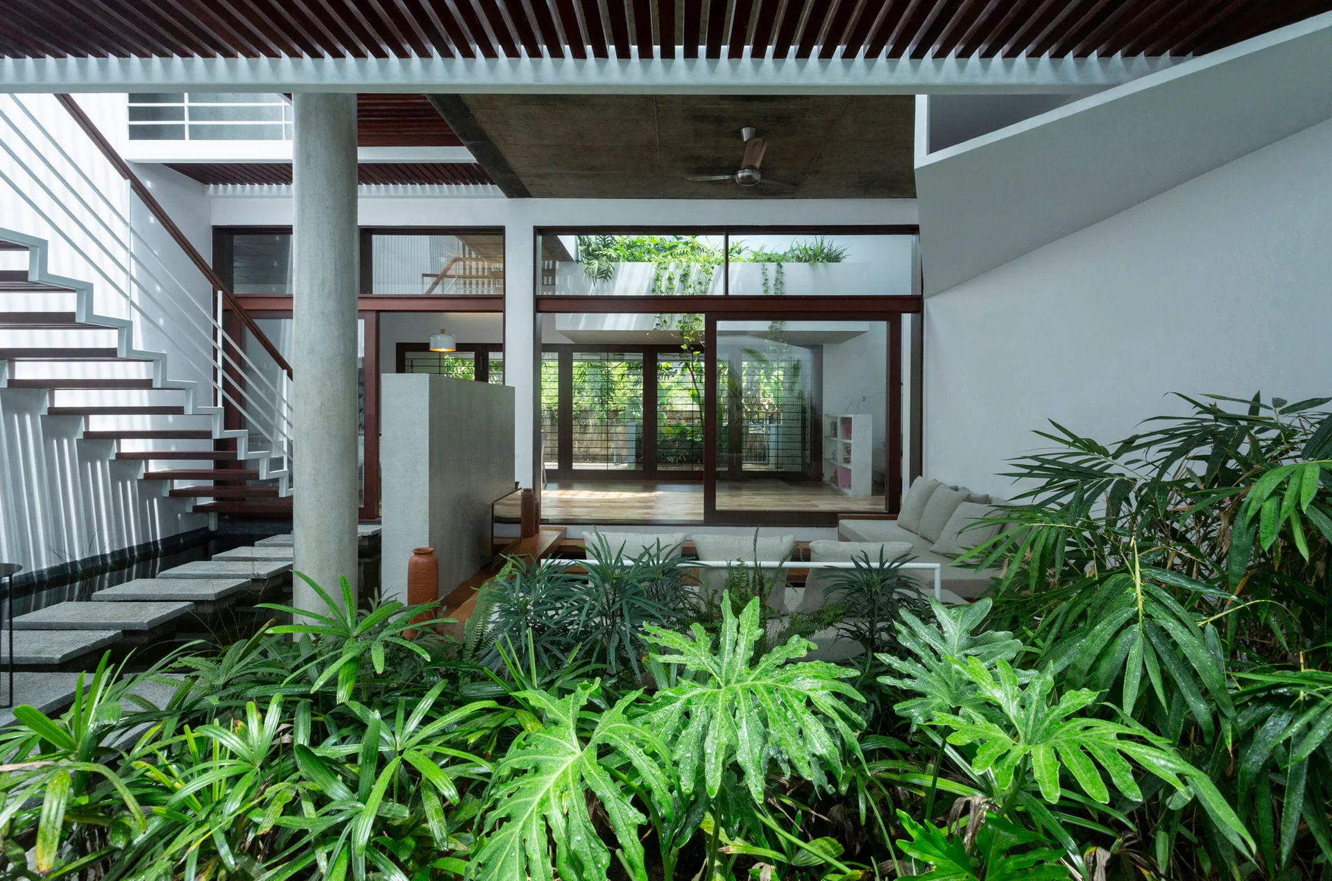 Modern interior courtyard featuring a stone stepping path over a pond next to a floating staircase and lush indoor foliage.