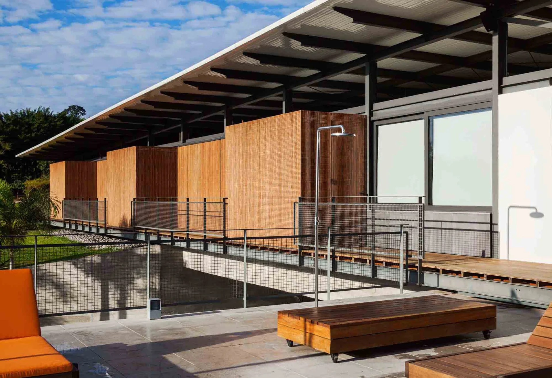 A modern outdoor deck features a slatted wood exterior, a stainless steel shower fixture, and a wooden bench on a tiled patio.