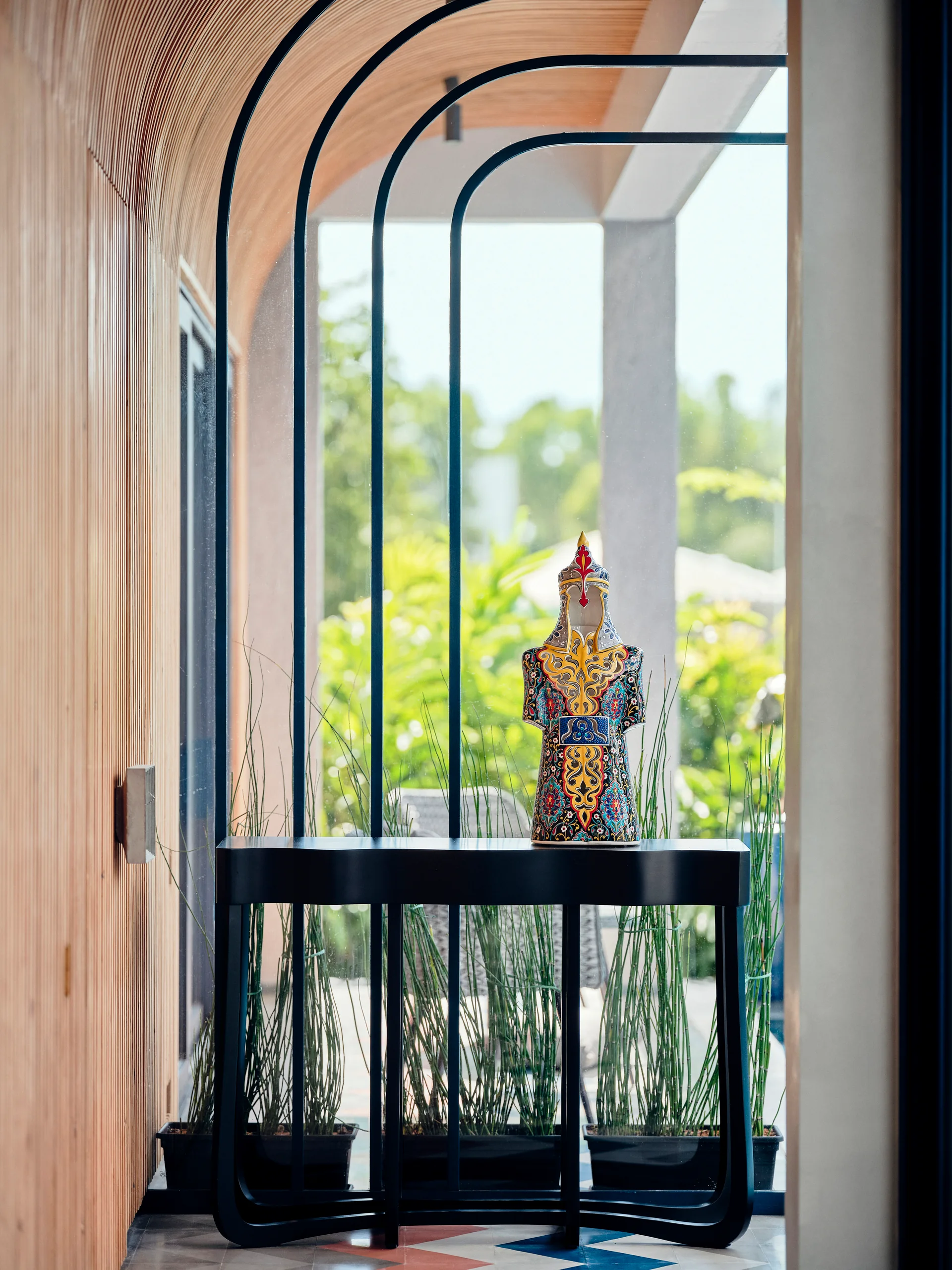 A black arched console table with an ornate colorful figurine sits in a hallway lined with wood slat walls in front of a glass window.
