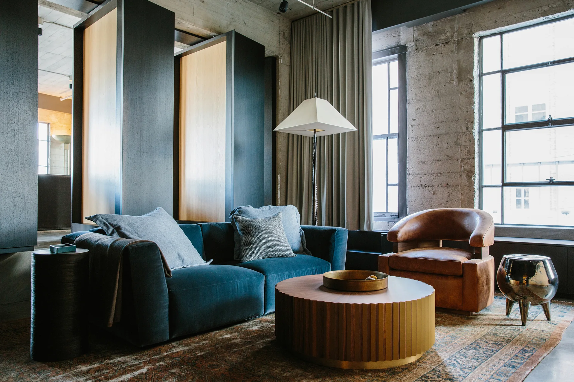 An industrial loft living room with a blue velvet sofa, a round ribbed wood coffee table, a brown leather armchair, and exposed concrete walls.