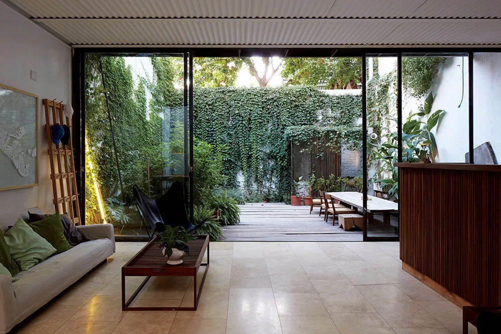 A modern living room with beige stone flooring and a grey sofa opens via large sliding glass doors to an outdoor courtyard surrounded by ivy-covered walls.