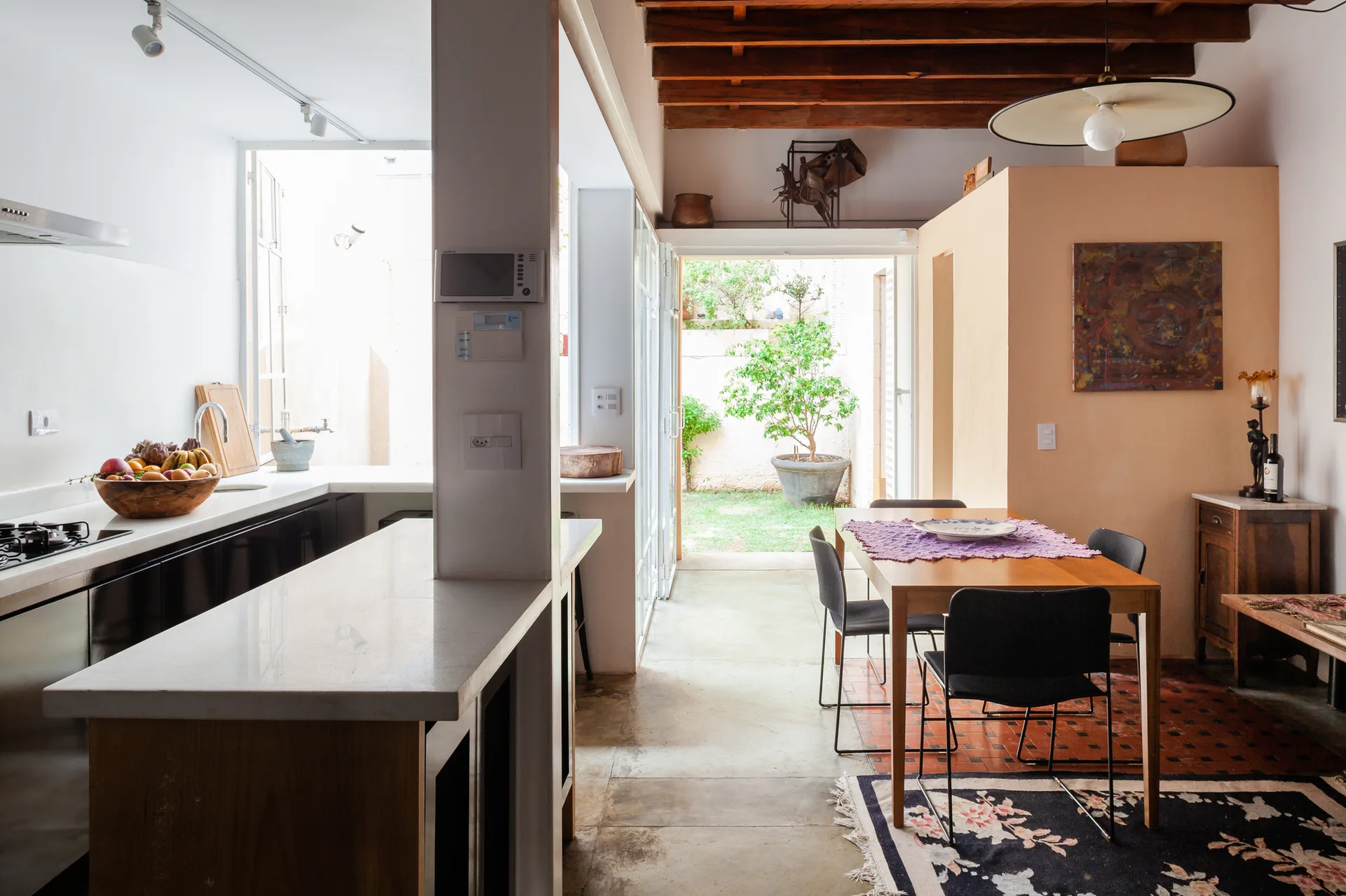 An open kitchen and dining space with a wooden dining table under exposed ceiling beams next to a doorway leading to a green patio.