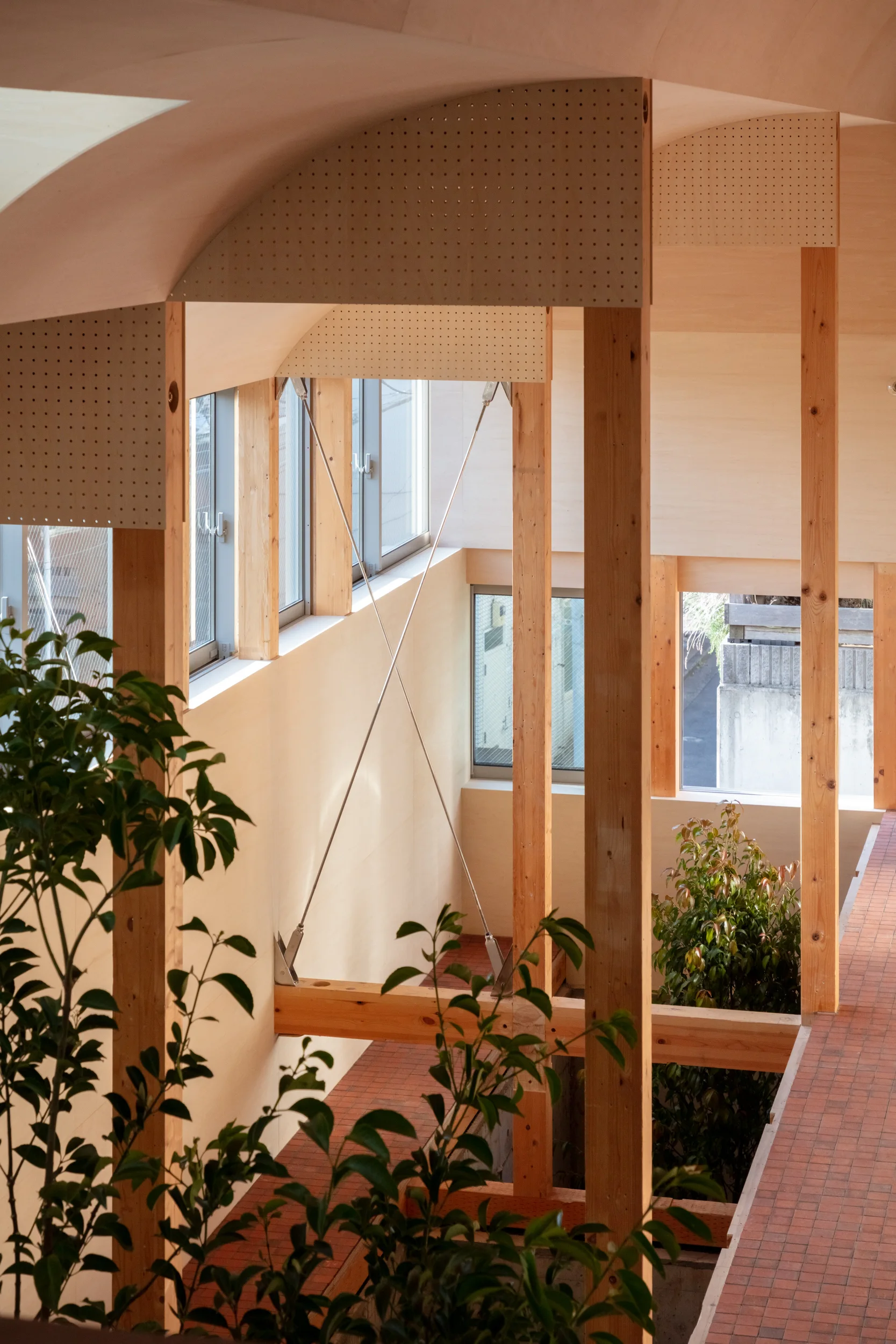 An interior architectural space features exposed timber posts, a curved wood-paneled ceiling, metal cross-bracing, and a terracotta tiled floor.