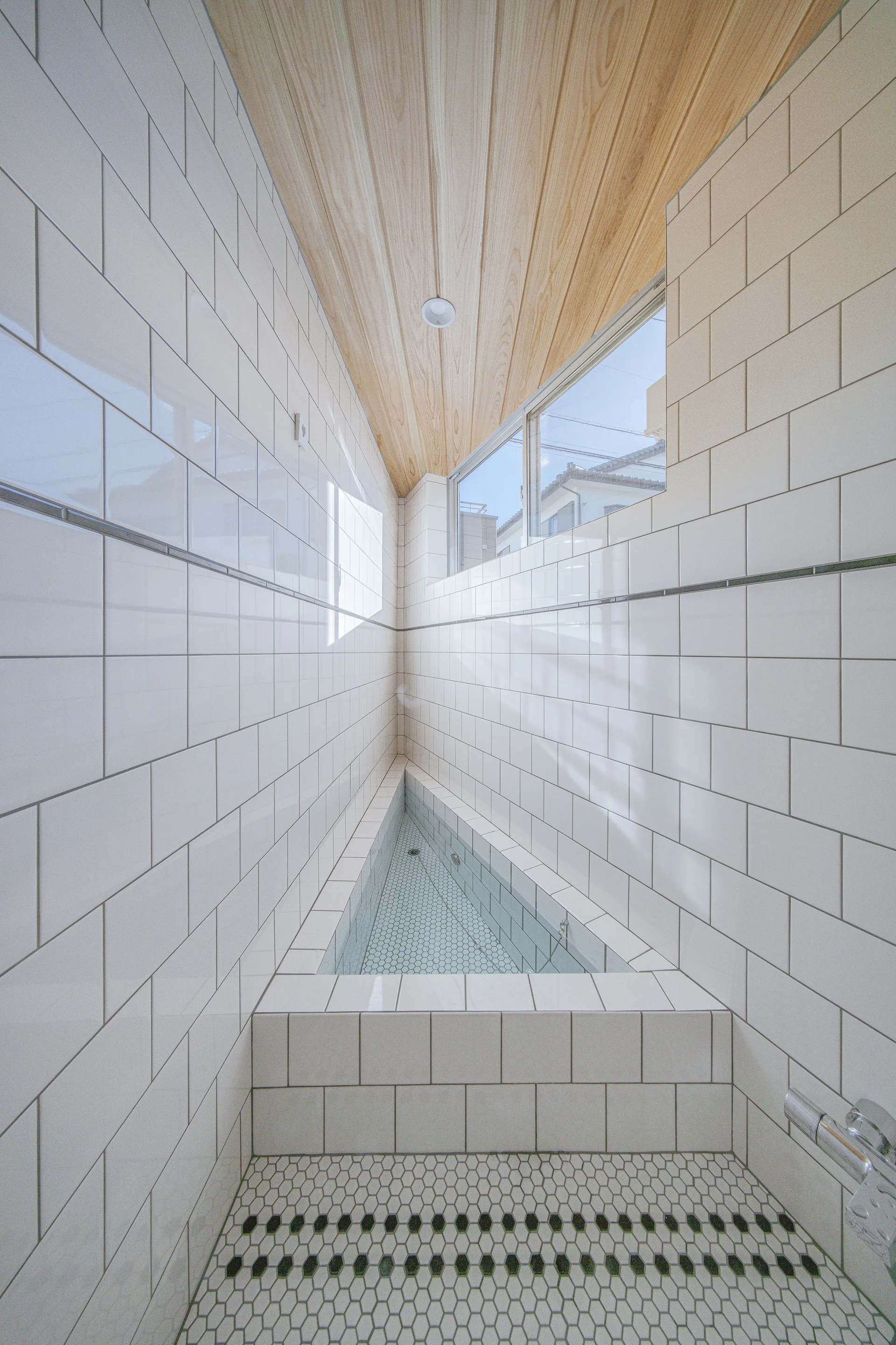 A narrow bathroom features a triangular built-in bathtub surrounded by white subway tile walls, hex tile flooring, and a wood plank ceiling.