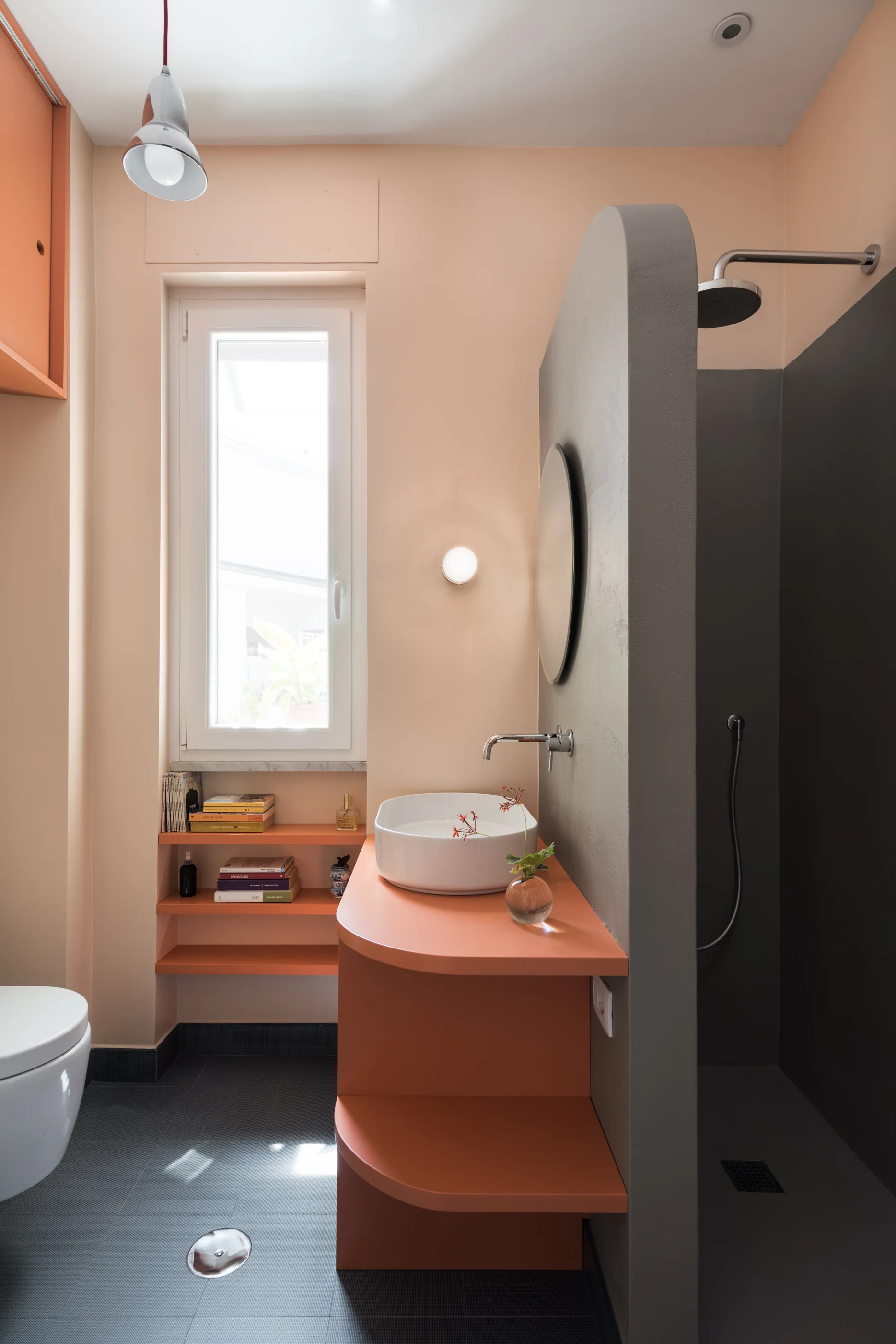 A modern peach bathroom featuring a curved orange vanity, a white vessel sink, a grey plaster shower partition, and dark grey tile floors.