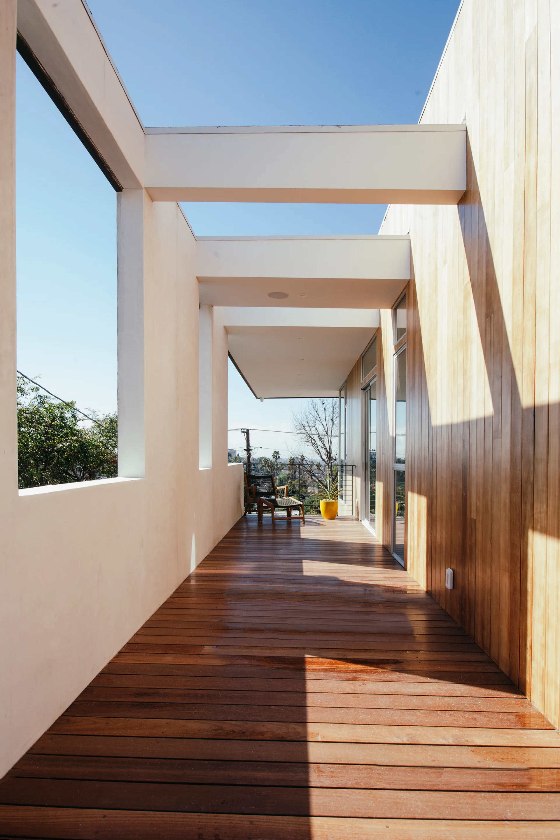 A modern outdoor balcony features warm wood plank decking, matching vertical wood wall paneling, and white structural beams framing a clear blue sky.