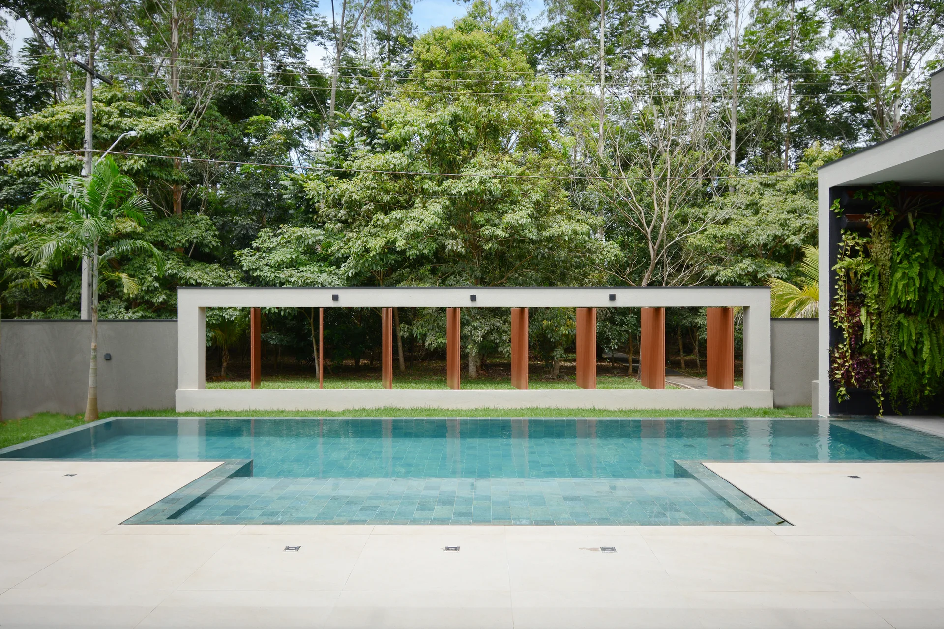 A rectangular swimming pool surrounded by a light beige patio, with a concrete screen wall featuring wood-toned slats set against a backdrop of trees.