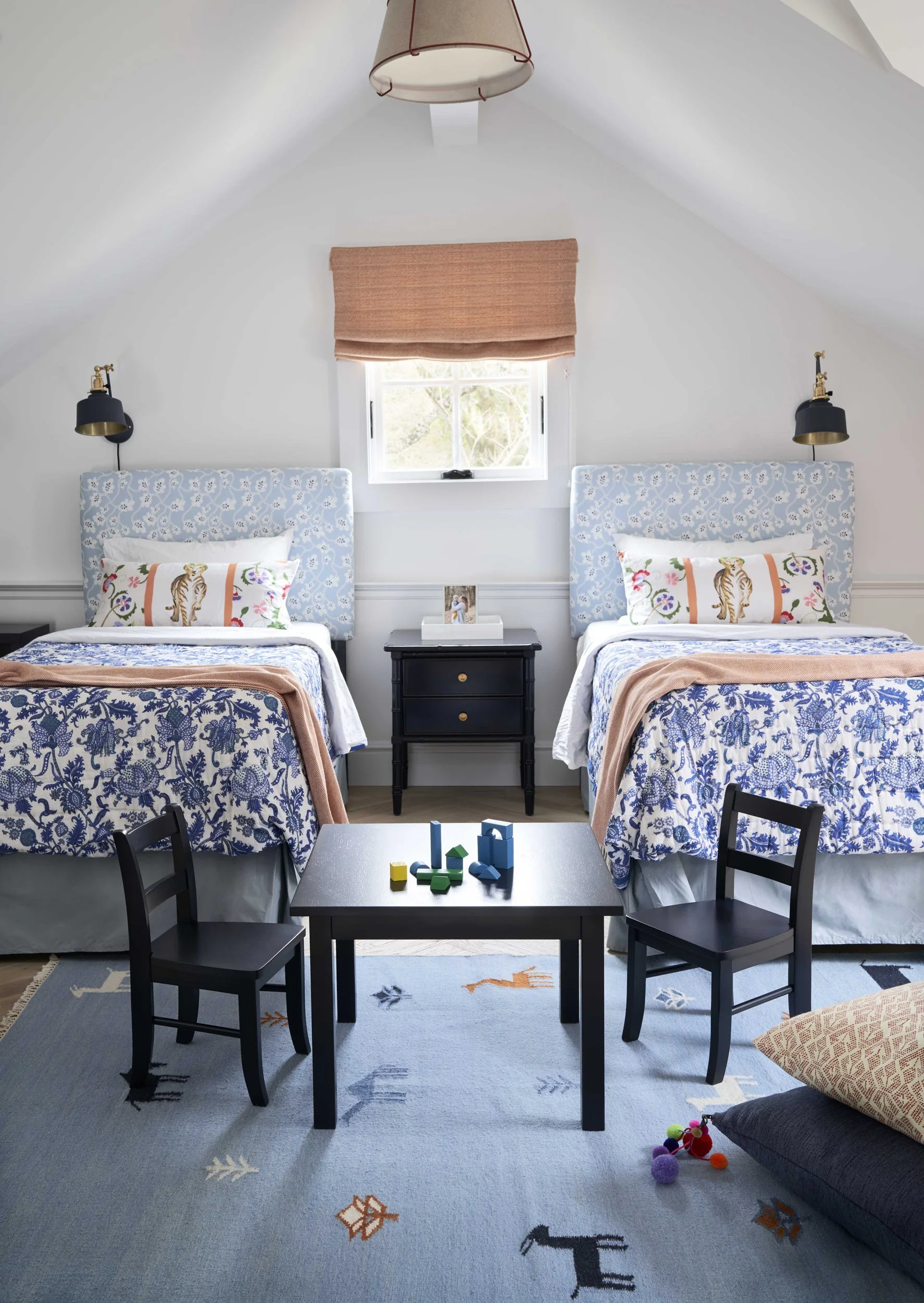 A symmetrical attic kids' bedroom with two twin beds featuring patterned blue floral headboards, a central black play table, and a blue patterned rug.