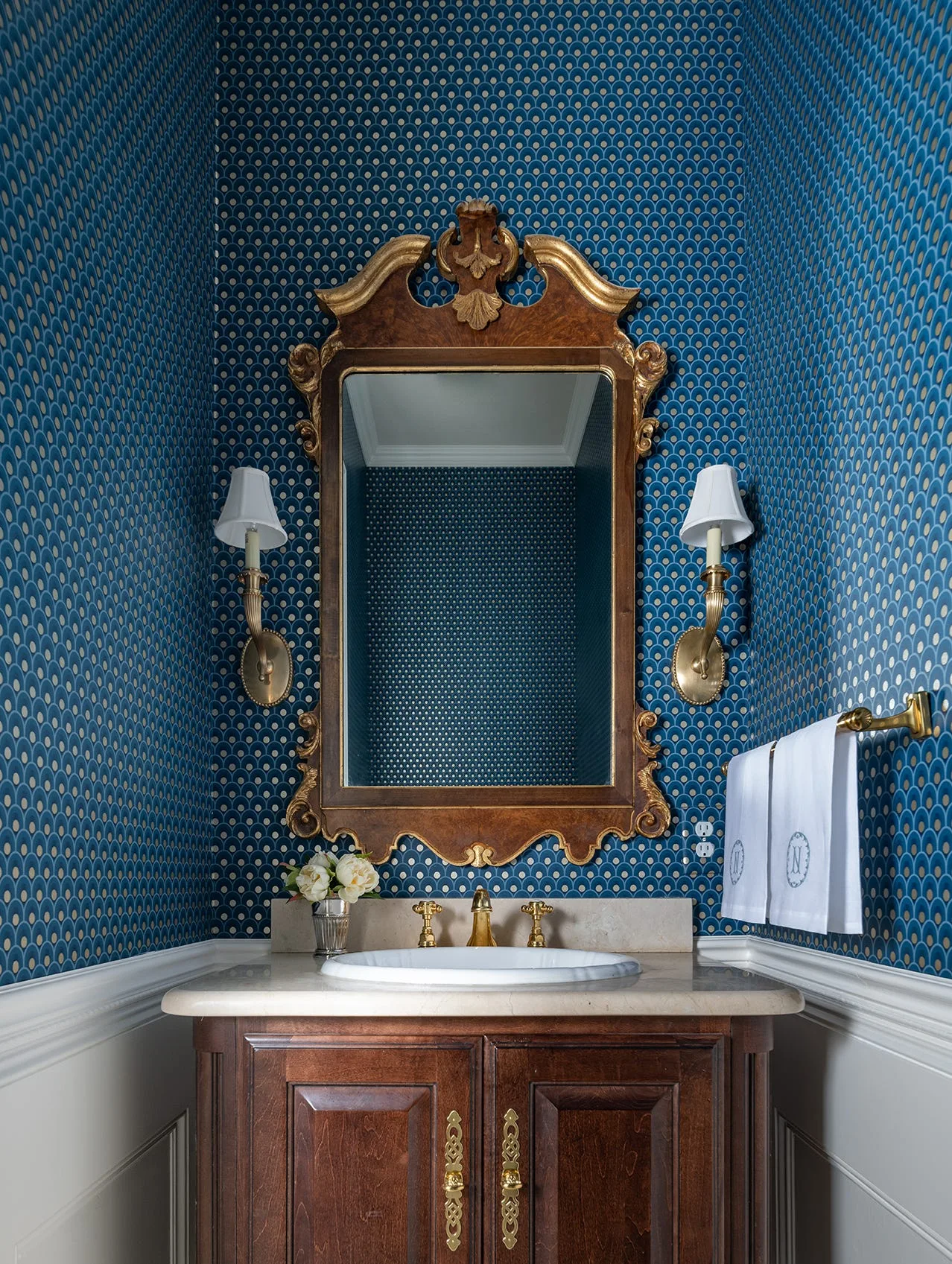 A powder room features a dark wood vanity, white wainscoting, blue geometric wallpaper, and an ornate gold-framed mirror flanked by brass sconces.
