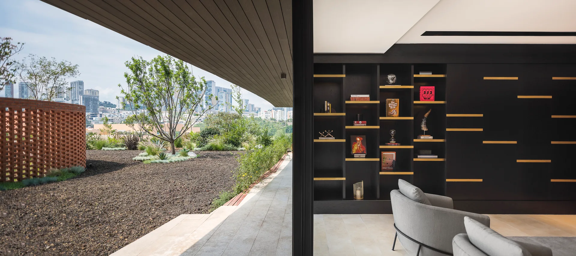 Modern living room featuring black integrated shelving and grey armchairs opening onto an outdoor patio with a curved brick wall and city skyline view.