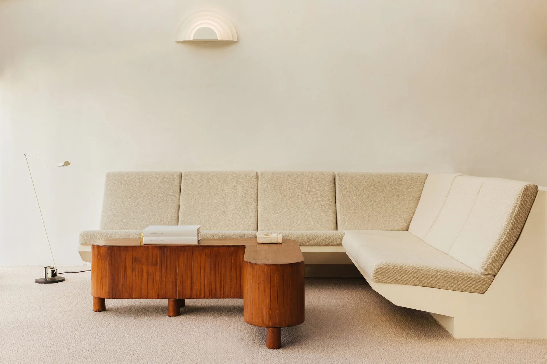 A cream sectional sofa sits against an off-white plaster wall next to a curved wood coffee table and an arched wall sconce.