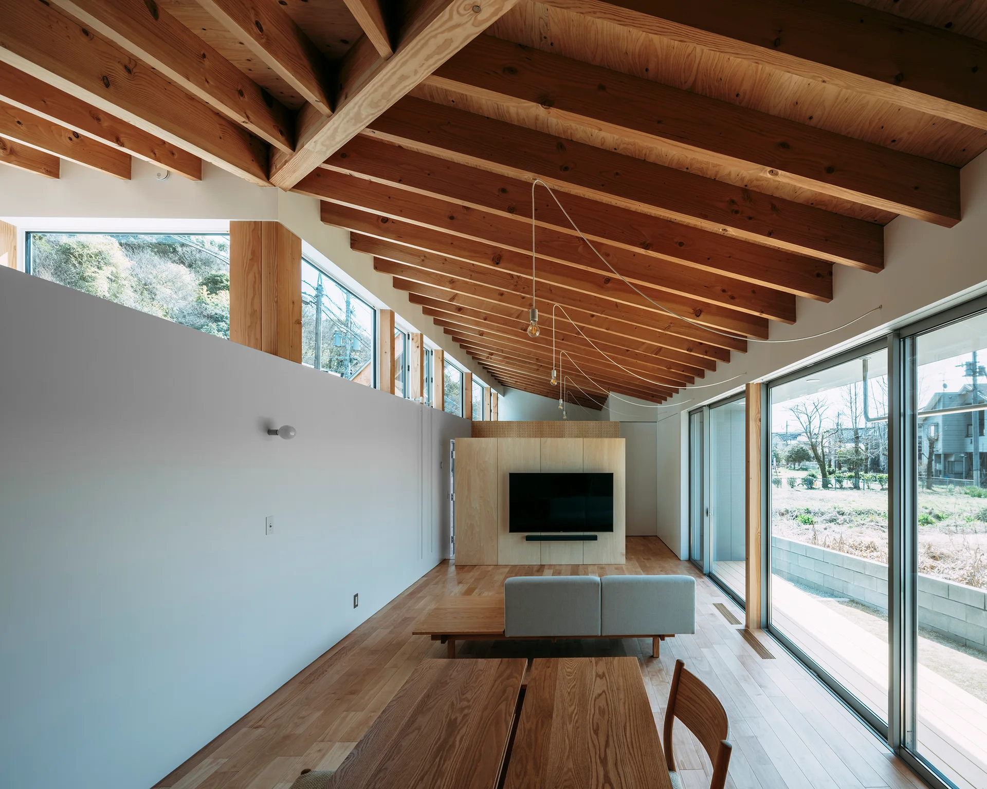 A minimalist living room features a vaulted wood ceiling with exposed rafters, light wood floors, a grey sofa, and large sliding glass doors.