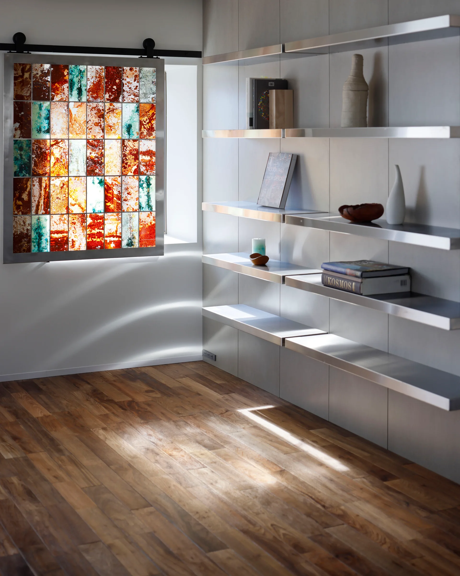 A sliding stained glass window panel hangs on a black track next to minimalist metal shelves on a gray paneled wall over hardwood flooring.