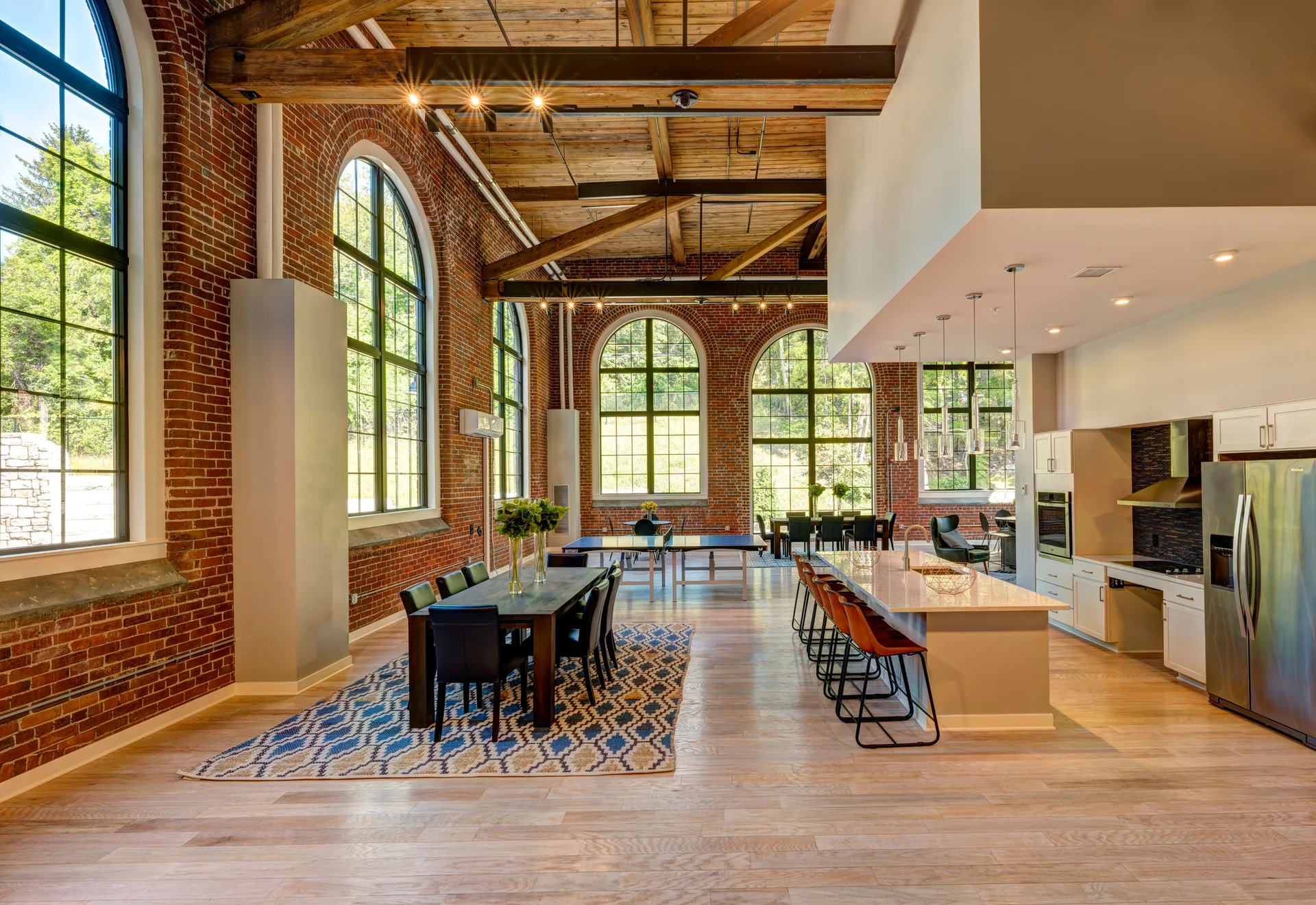 An open-plan industrial loft interior featuring exposed brick walls, large arched windows, a wood plank ceiling, a dining set, and a modern kitchen.