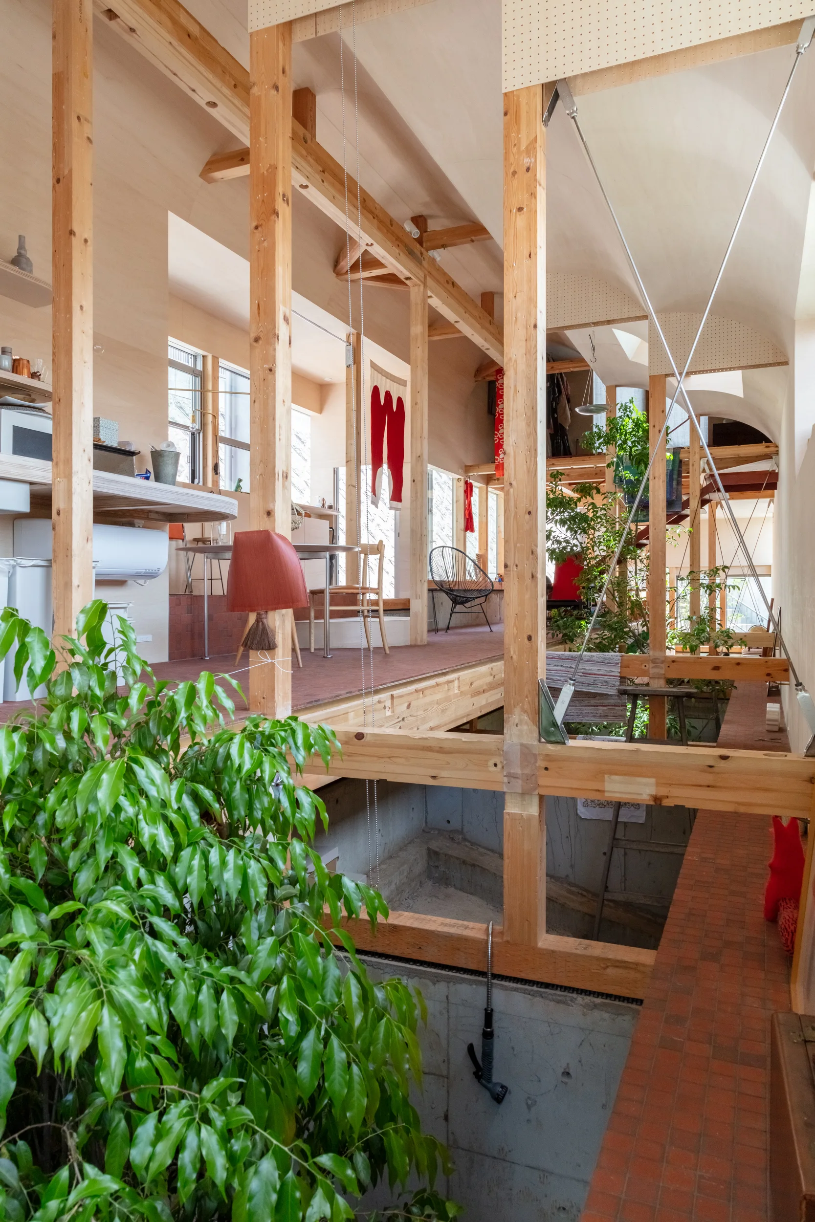 A multi-level residential interior featuring an exposed timber frame structure, terracotta tile flooring, concrete foundation walls, and green potted plants.