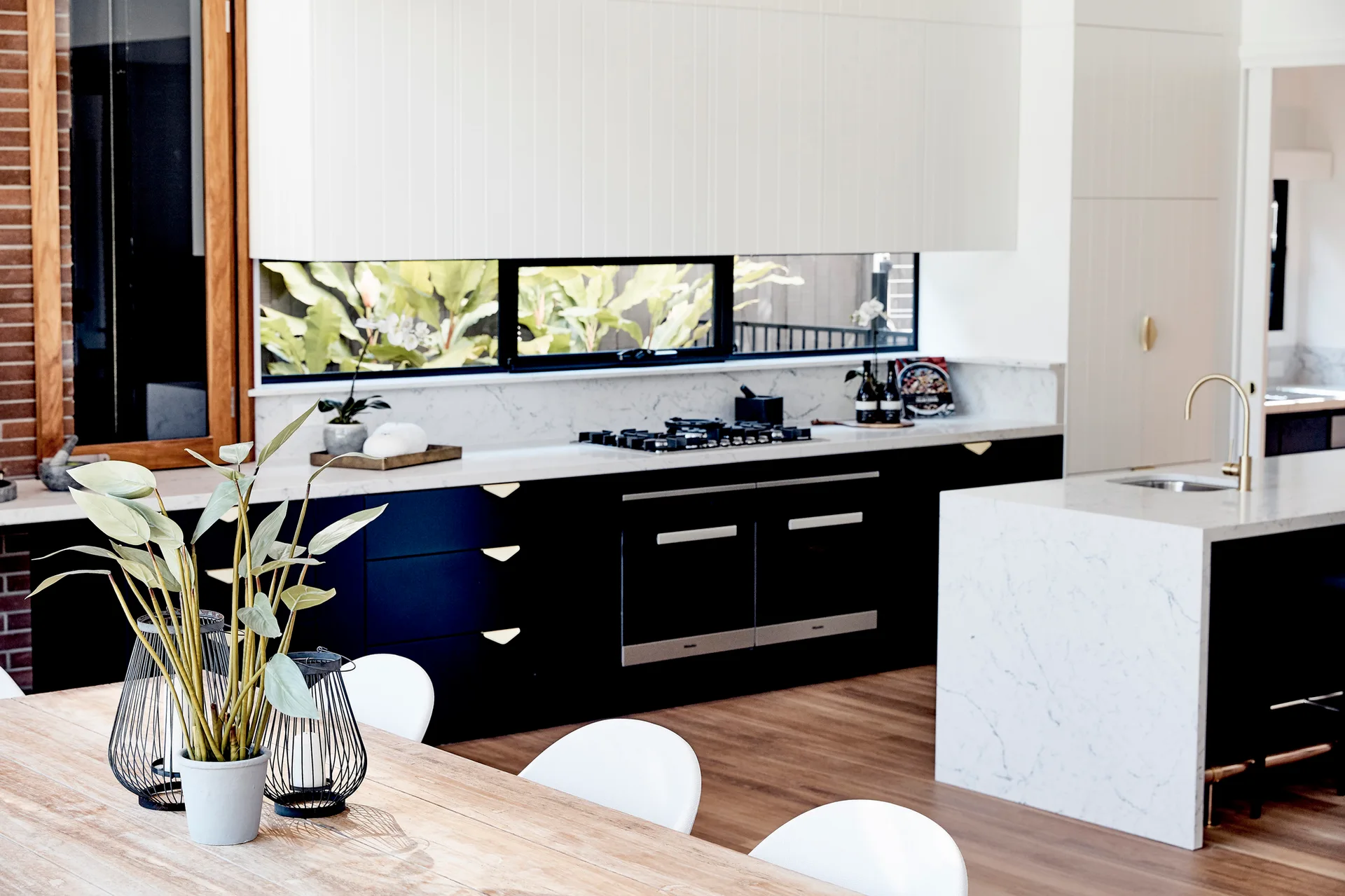 A modern kitchen featuring black lower cabinets, white upper cupboards, a marble island with a gold faucet, and a low horizontal window.