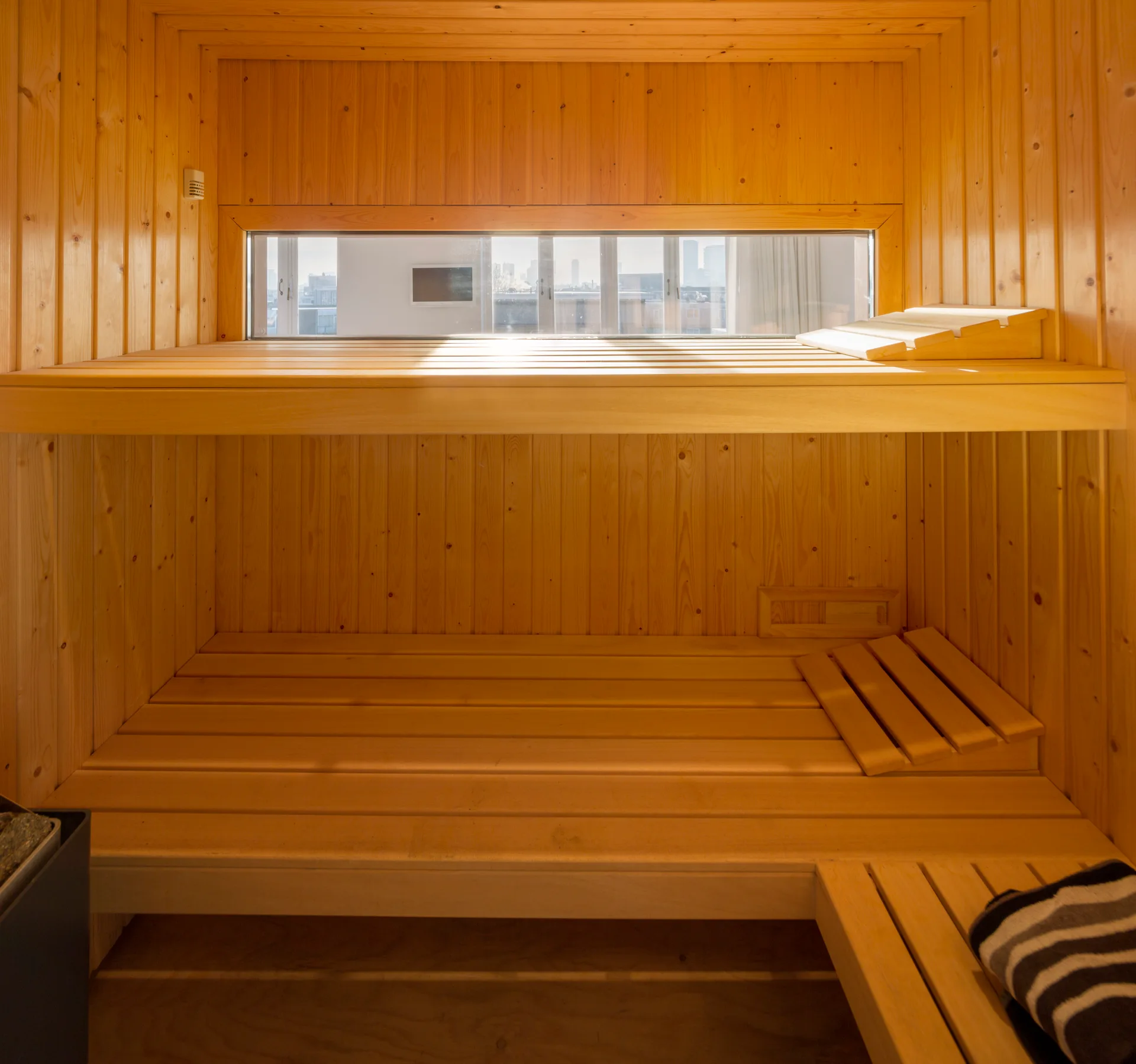 A natural wood sauna interior features tiered bench seating, vertical wall panels, and a narrow horizontal window overlooking a cityscape.