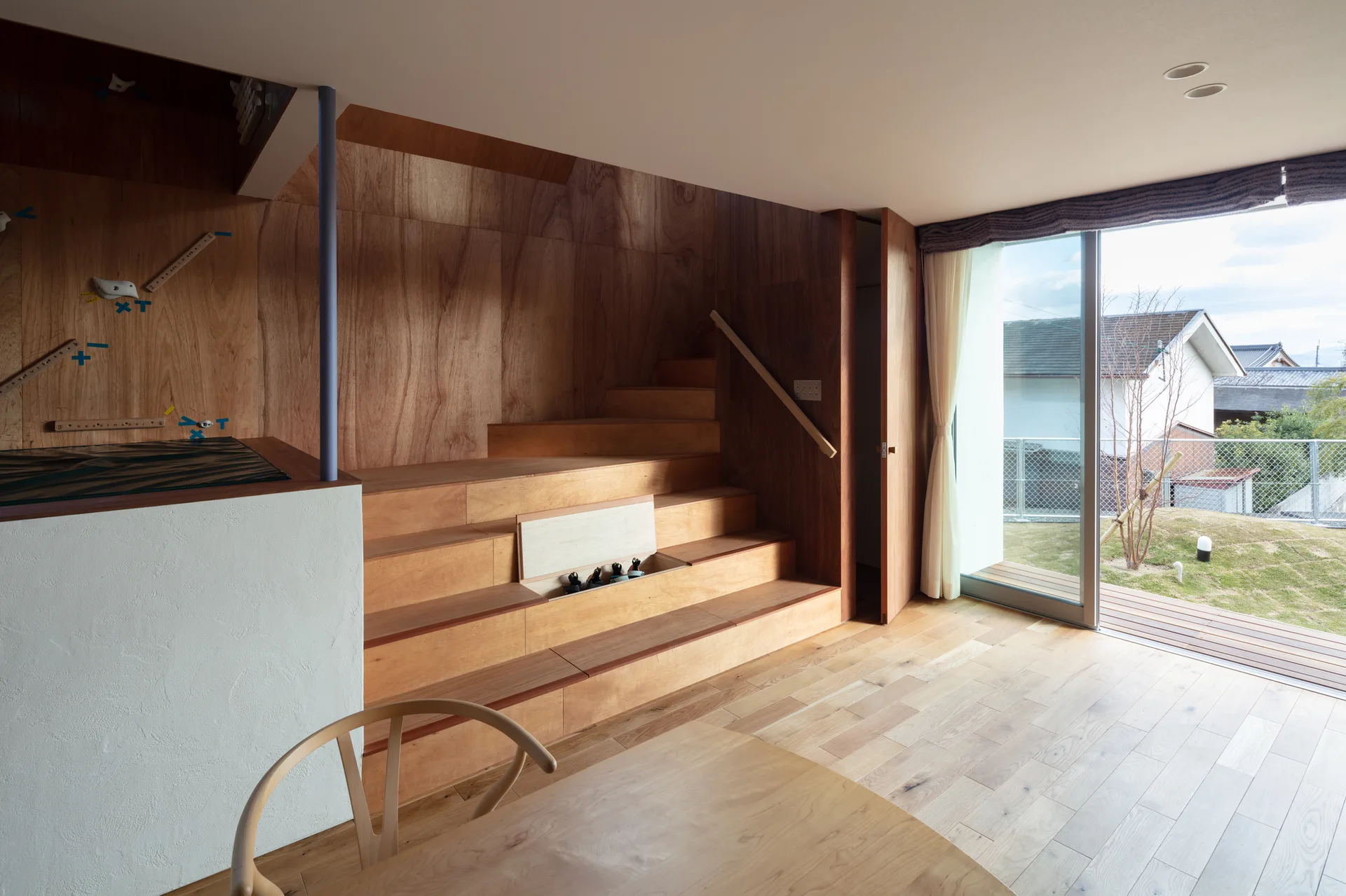 A minimalist wood paneled room features a wooden staircase with built-in storage compartments and sliding glass doors that open to a garden.