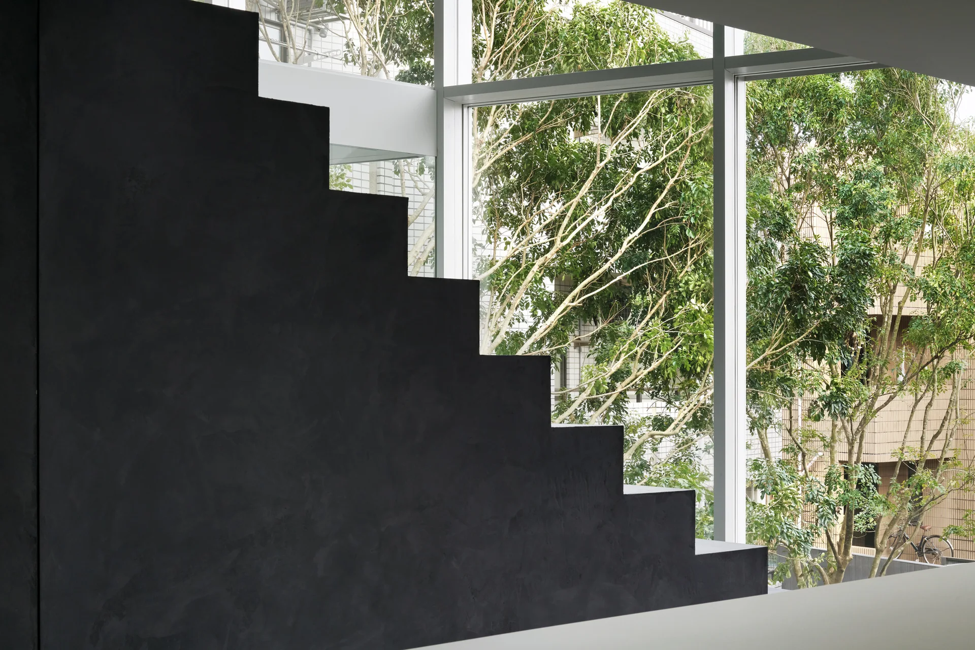 A dark matte black staircase sits in profile against a large glass window overlooking green trees outside.