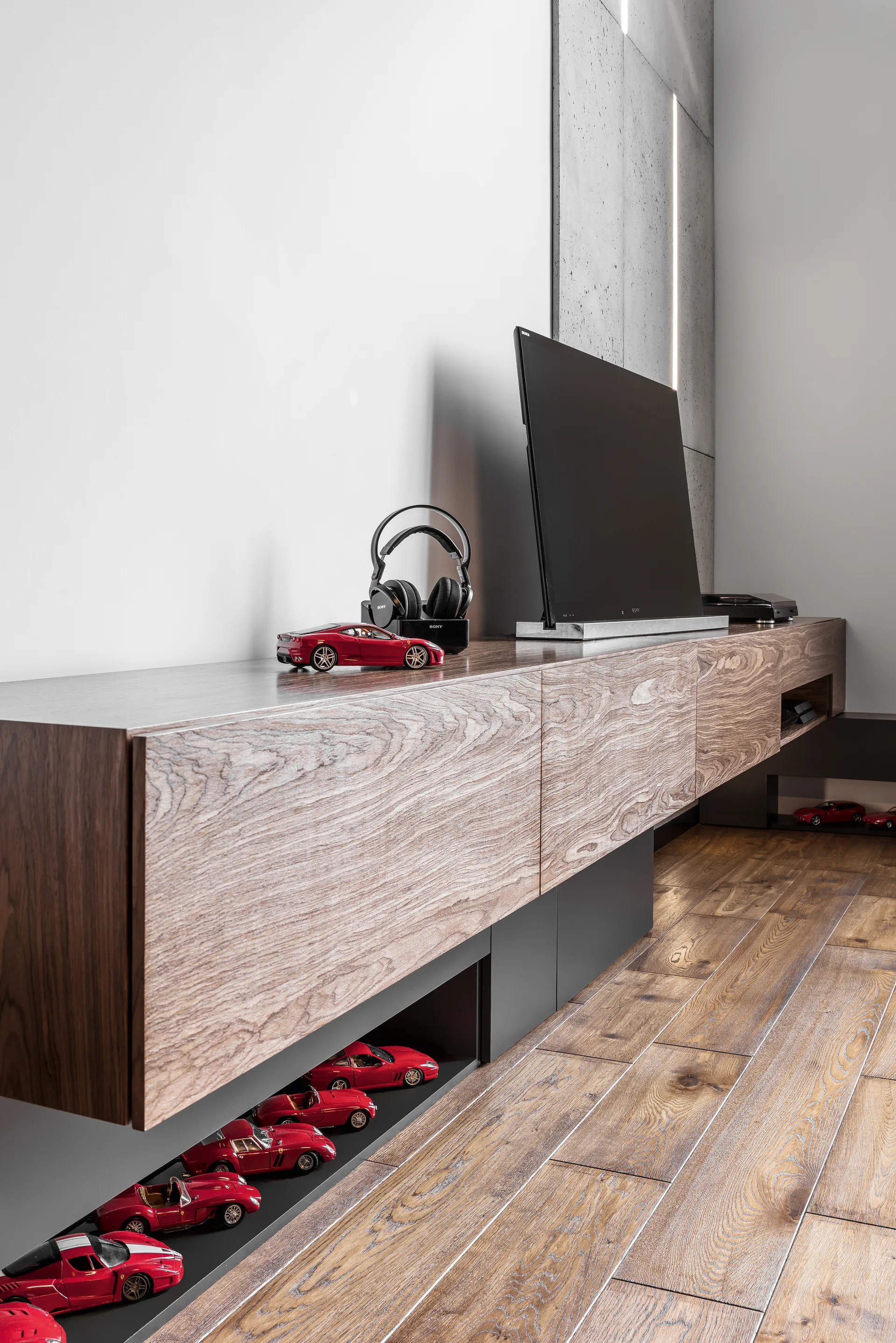 A wall-mounted wooden media console holds a television and headphones, while a lower shelf displays a collection of miniature red toy cars.