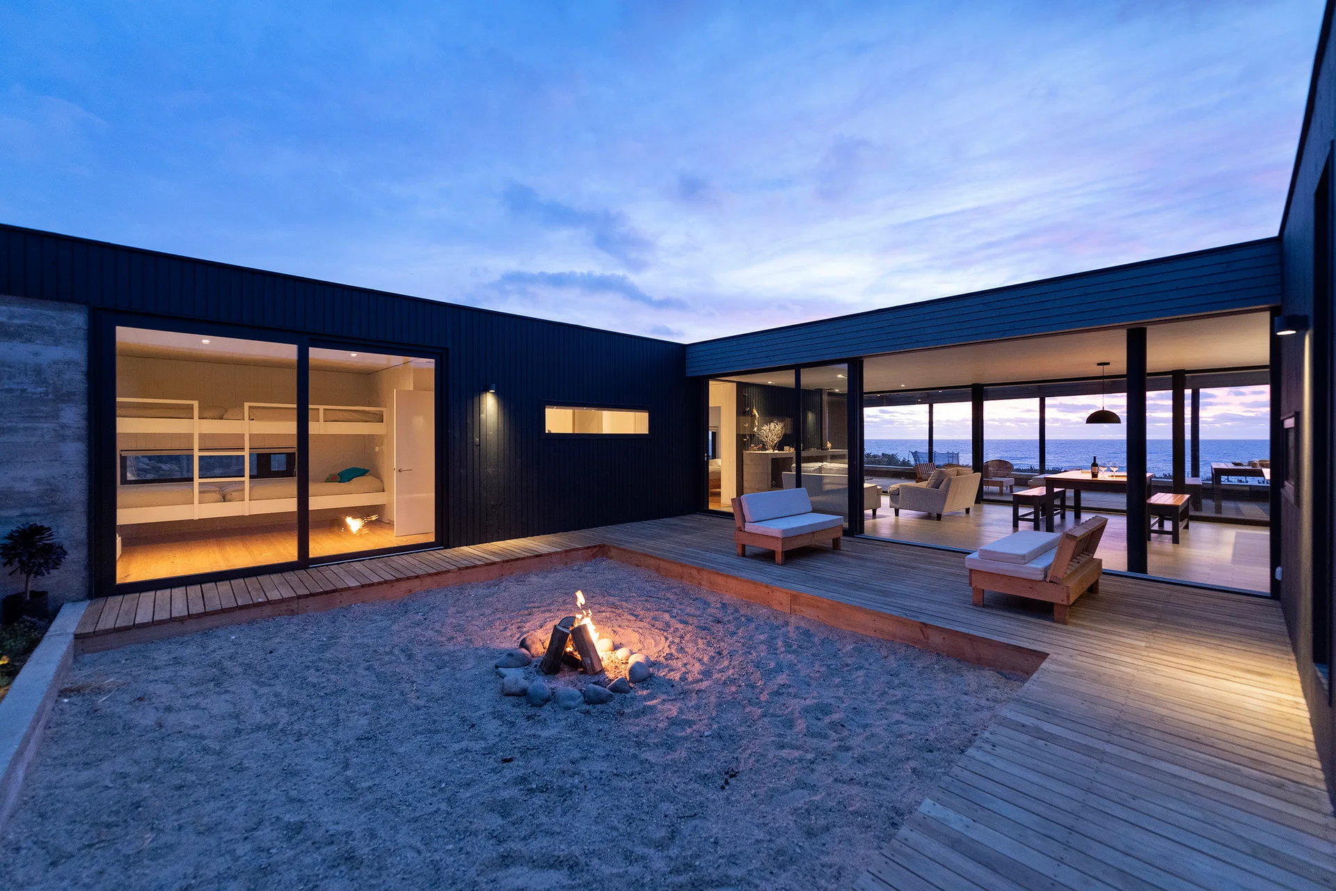 A modern coastal courtyard features a central fire pit in a sandy area surrounded by wood decking and glass-walled living spaces at dusk.