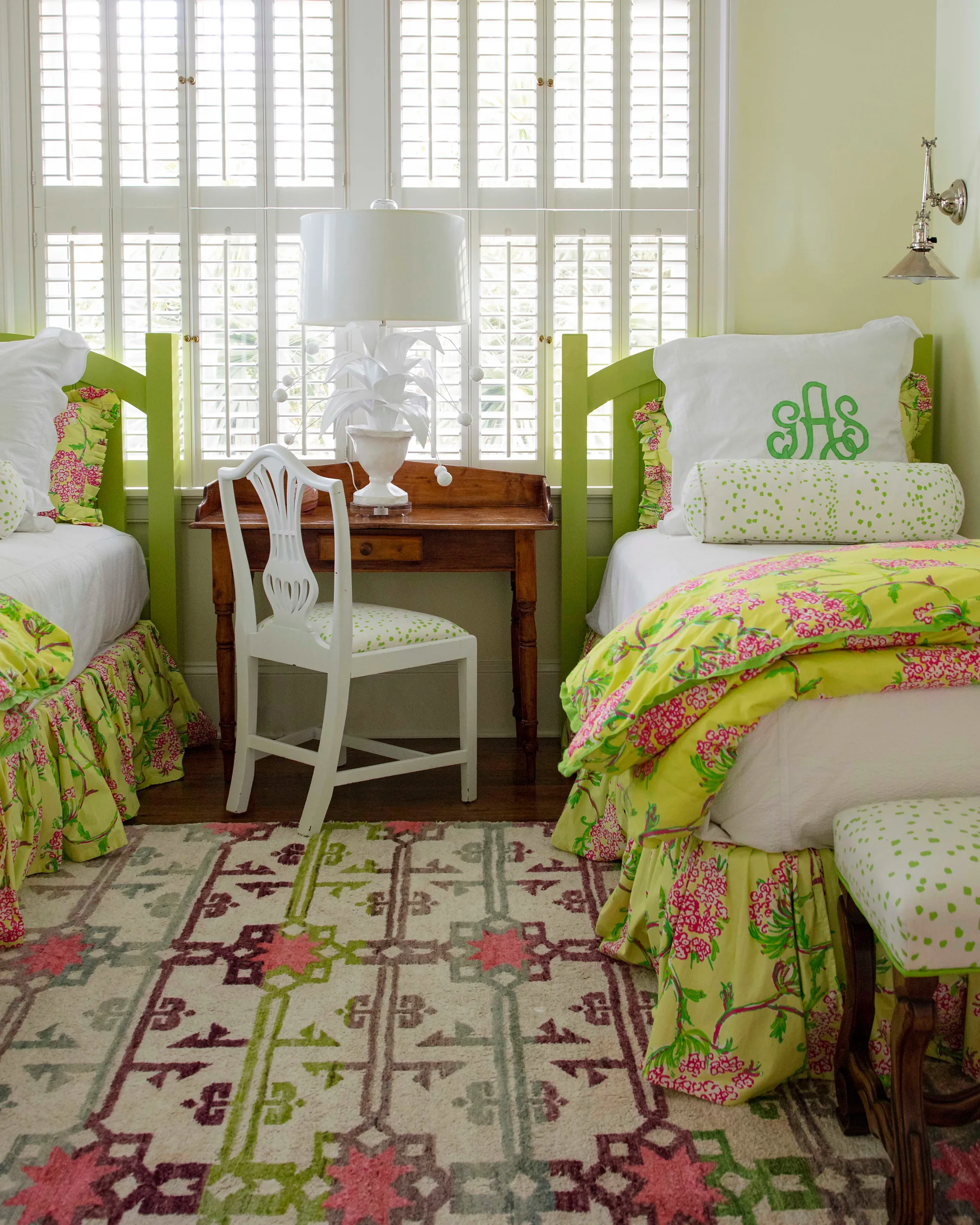 Two twin beds with green headboards and floral bedding flank a wooden writing desk in front of white shuttered windows.