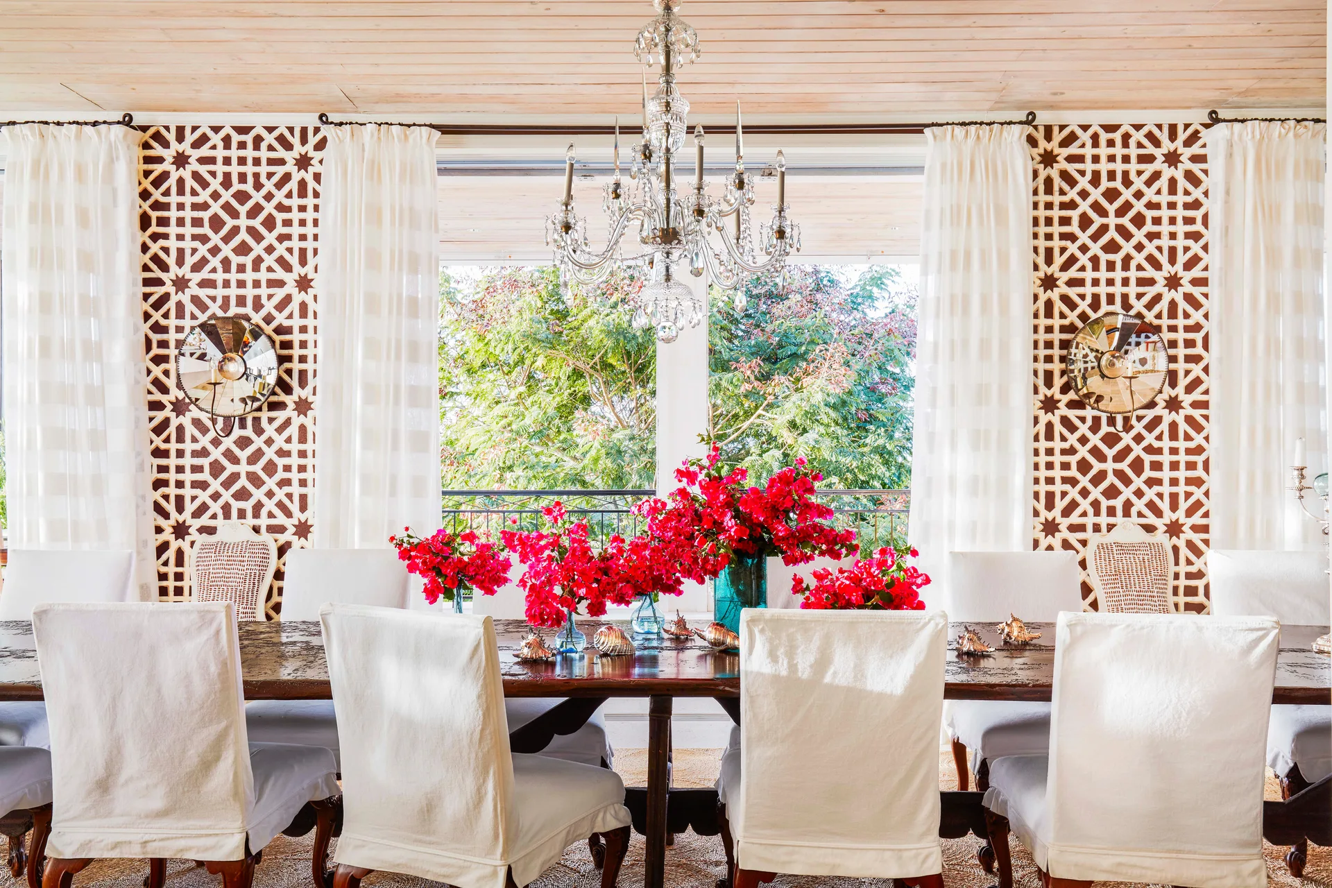 A formal dining room features a long wooden table with white slipcovered chairs under a crystal chandelier, flanked by brown geometric lattice walls.