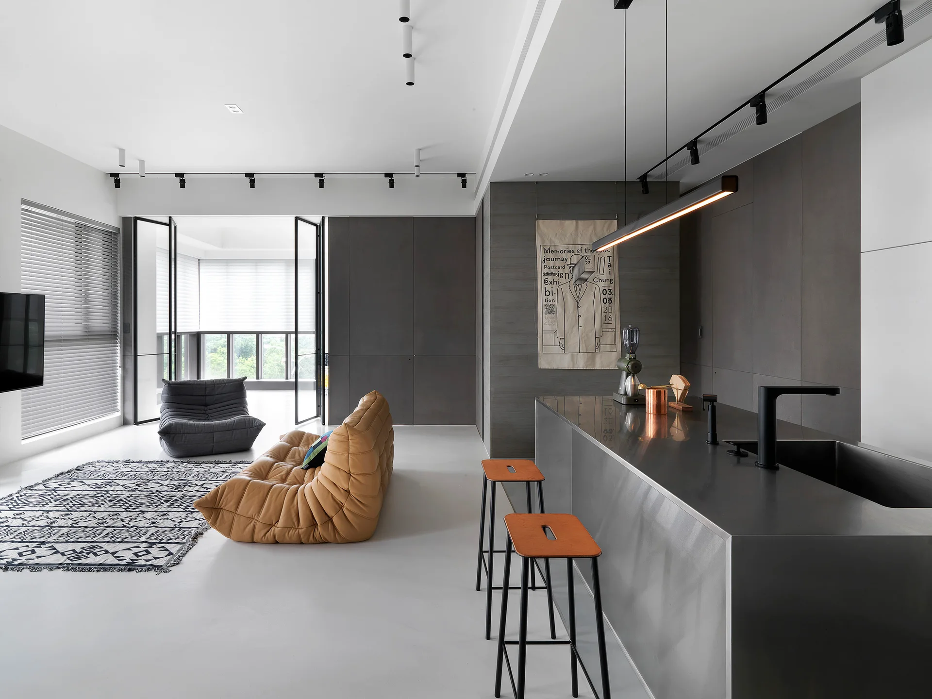 A modern open-plan kitchen and living space featuring a stainless steel island, two bar stools, a tan tufted sofa, and grey paneled walls.