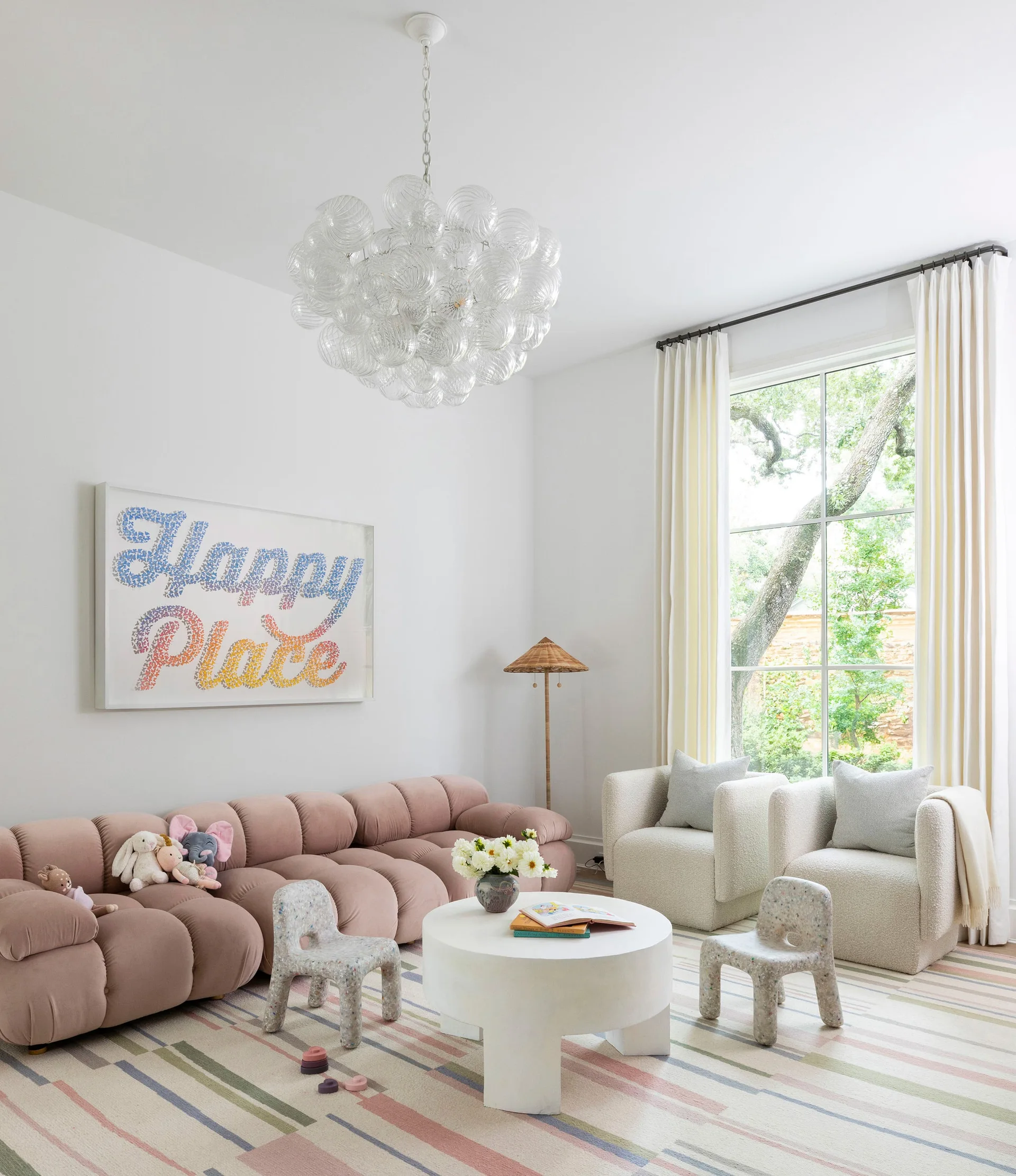 A contemporary playroom features a pink modular sofa, two white boucle armchairs, a striped rug, and a bubble glass chandelier under a high ceiling.
