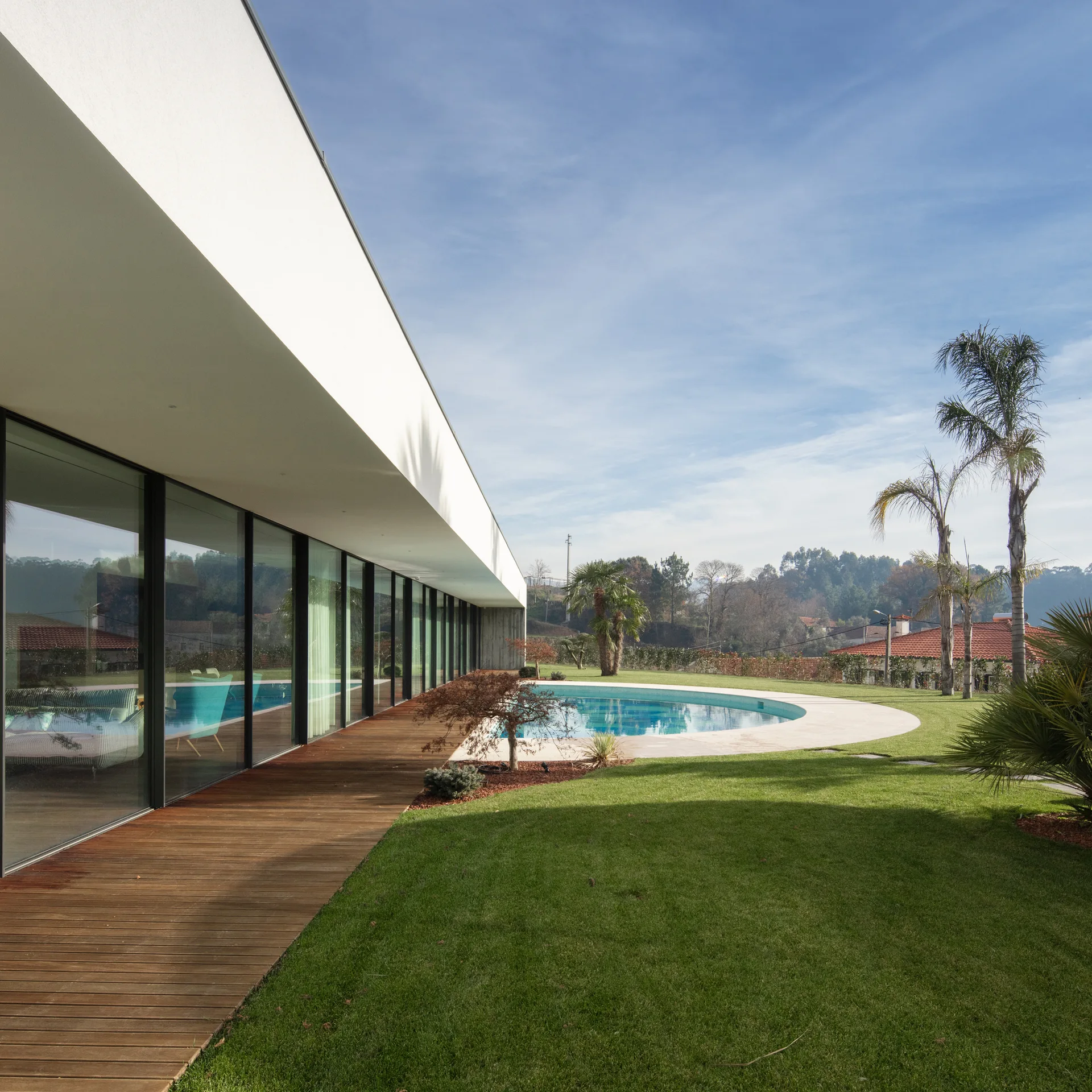A modern white home features a long glass facade, a wooden deck, and a curved swimming pool set within a green lawn.