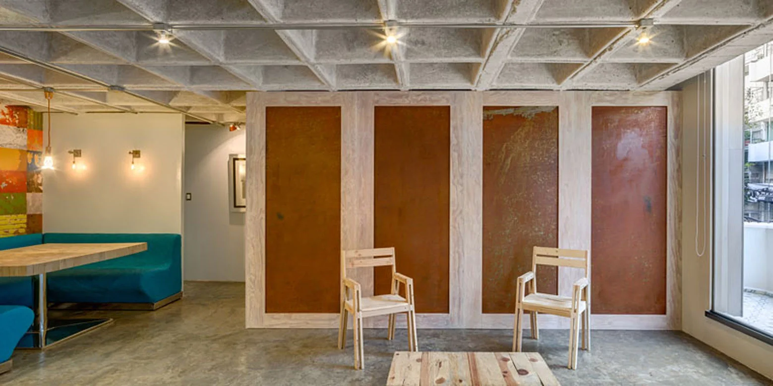 A modern living space features an exposed concrete waffle ceiling, teal booth seating, and a light wood partition wall with inset rusted metal panels.