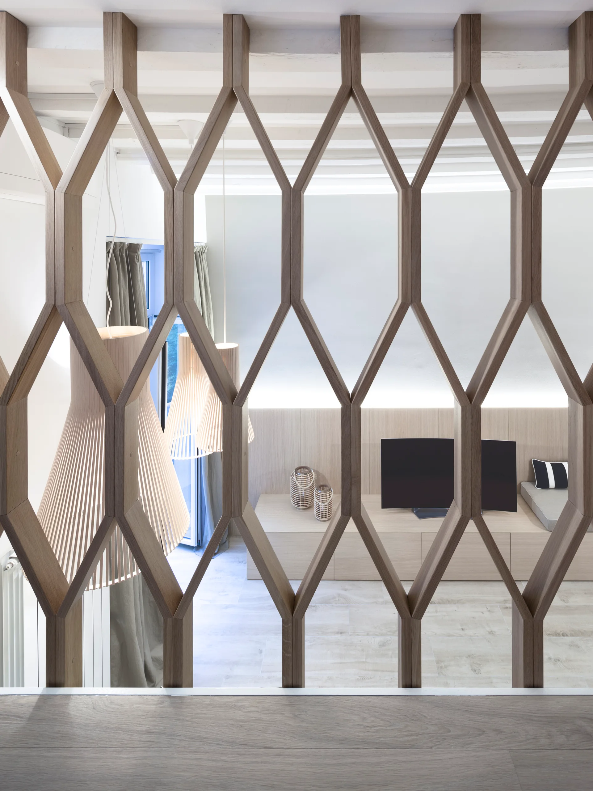 A geometric wood screen separates a modern living room featuring slatted pendant lights, a low-profile media console, and neutral tiled flooring.