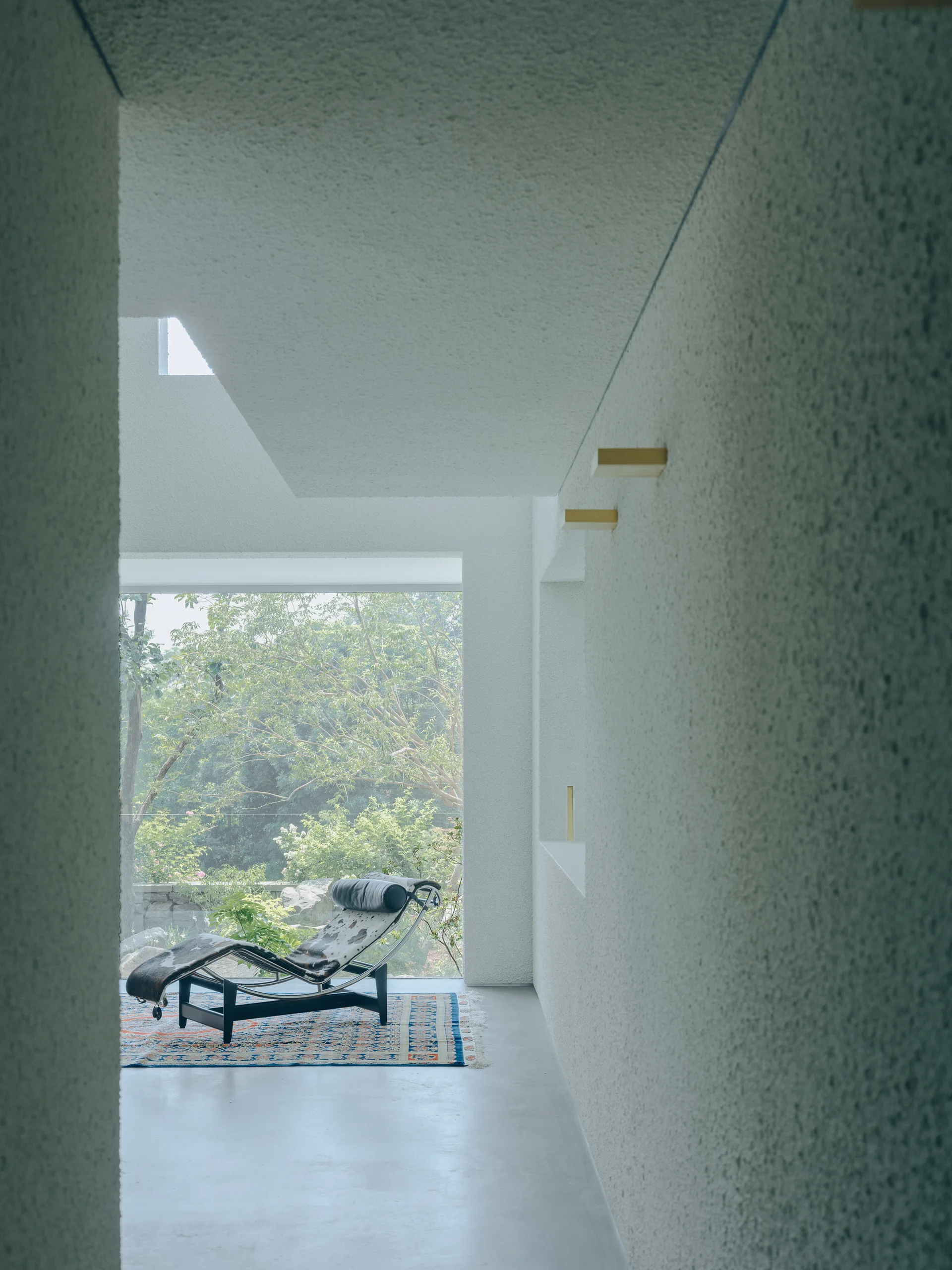 A modern chaise lounge rests on a patterned area rug in front of a large picture window, flanked by heavily textured plaster walls.