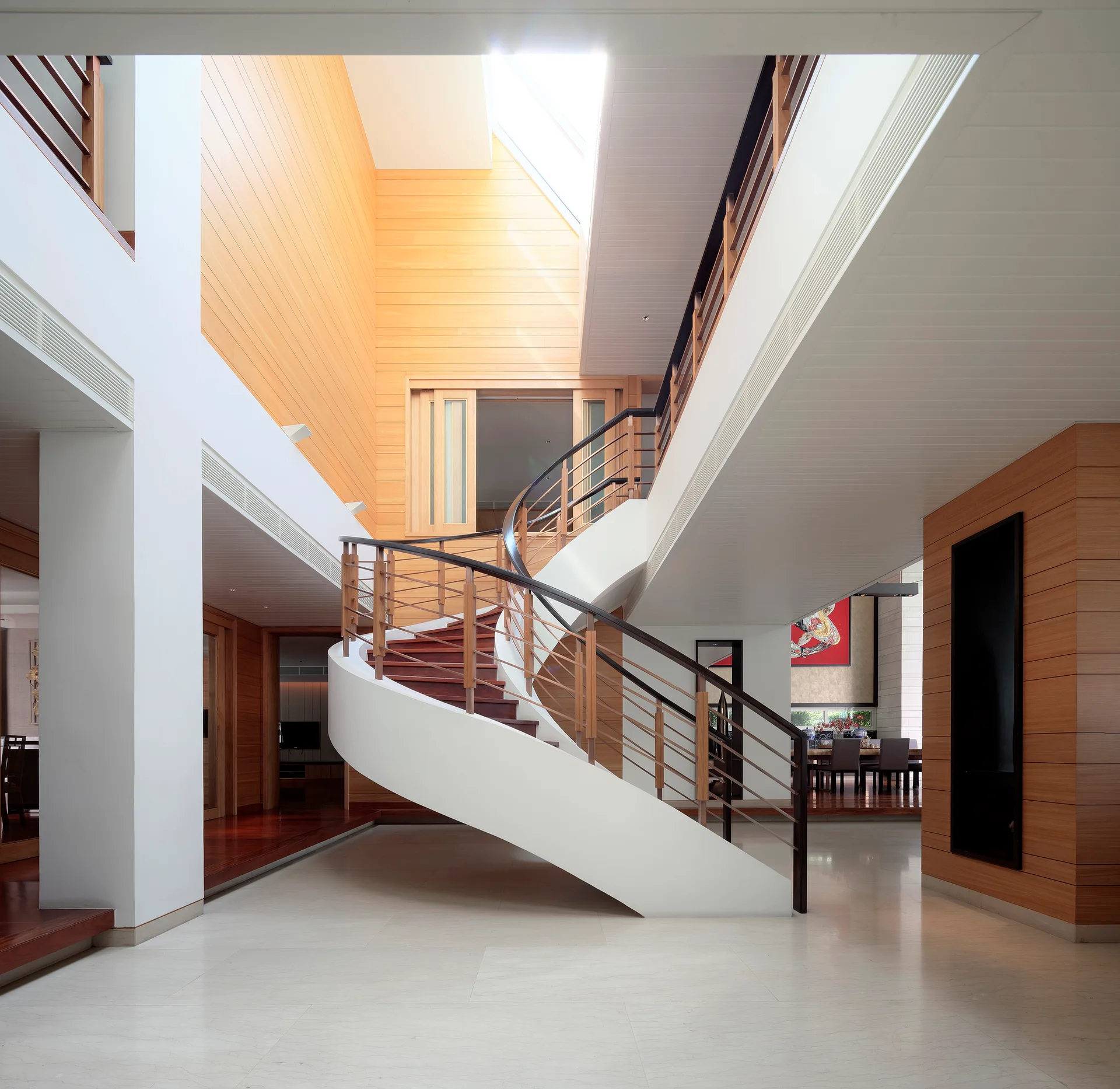 A modern atrium features a central white spiral staircase with wood treads, flanked by warm wood-paneled walls and a bright skylight above.