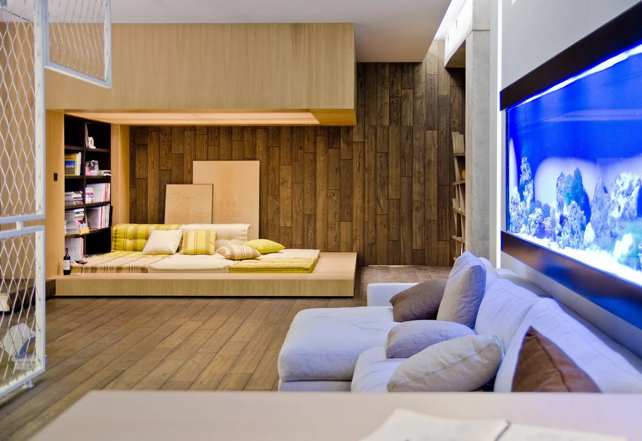 A contemporary living room featuring a white sectional sofa, a glowing blue wall-mounted aquarium, and a raised wooden lounge platform in the background.