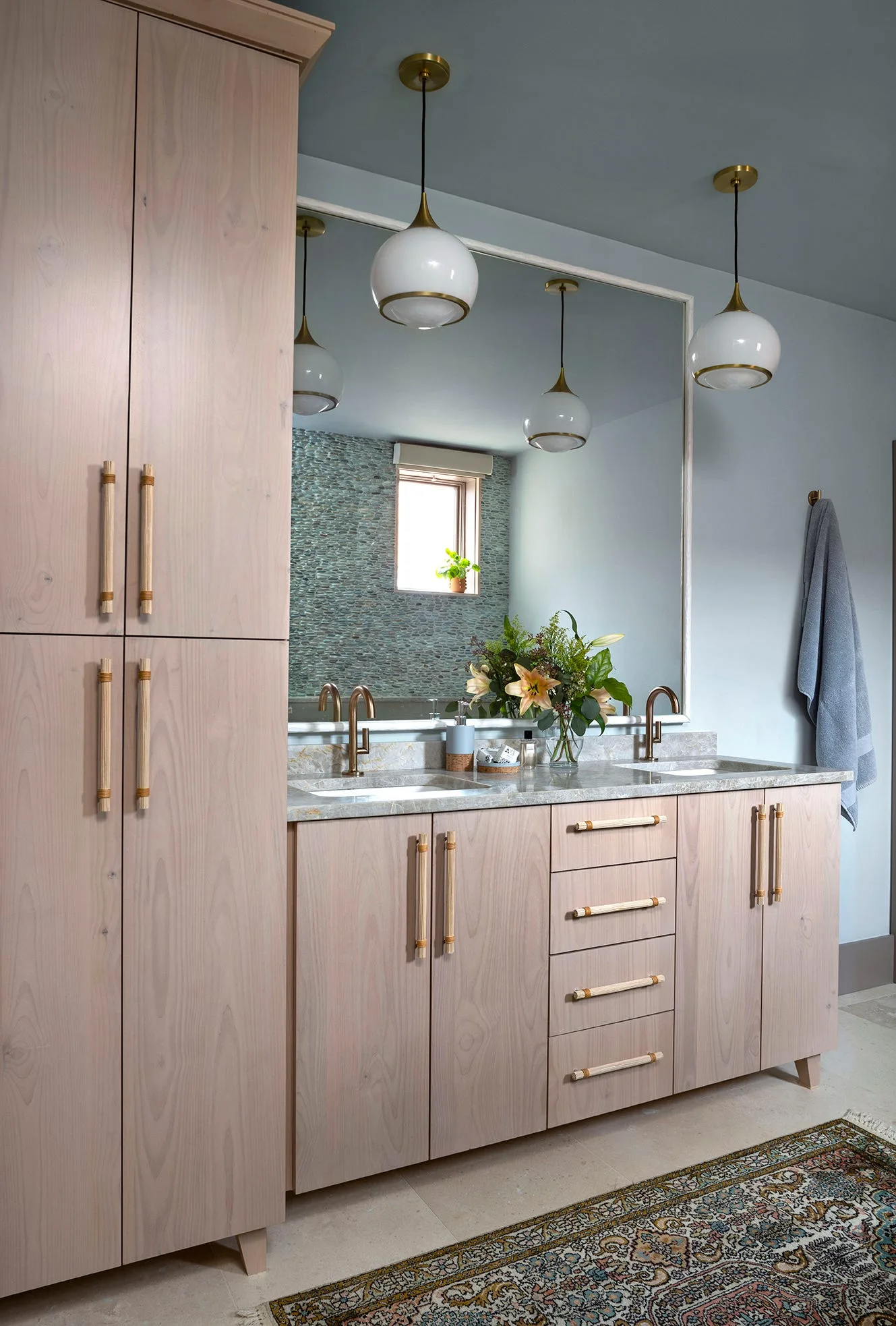 A bathroom features a light wood double vanity with brass hardware, globe pendant lights, and a large mirror reflecting a pebble-textured wall.